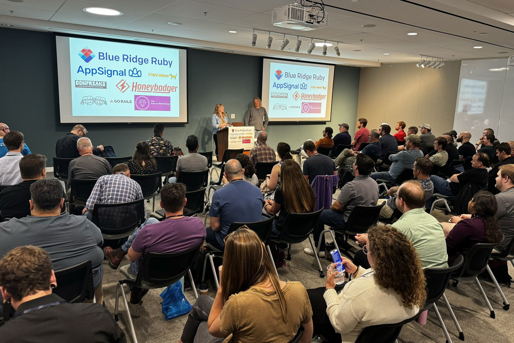 Two speakers addressing a large audience at the Blue Ridge Ruby conference, with sponsor banners including AppSignal and Honeybadger visible behind them, in a room filled with seated attendees listening attentively.