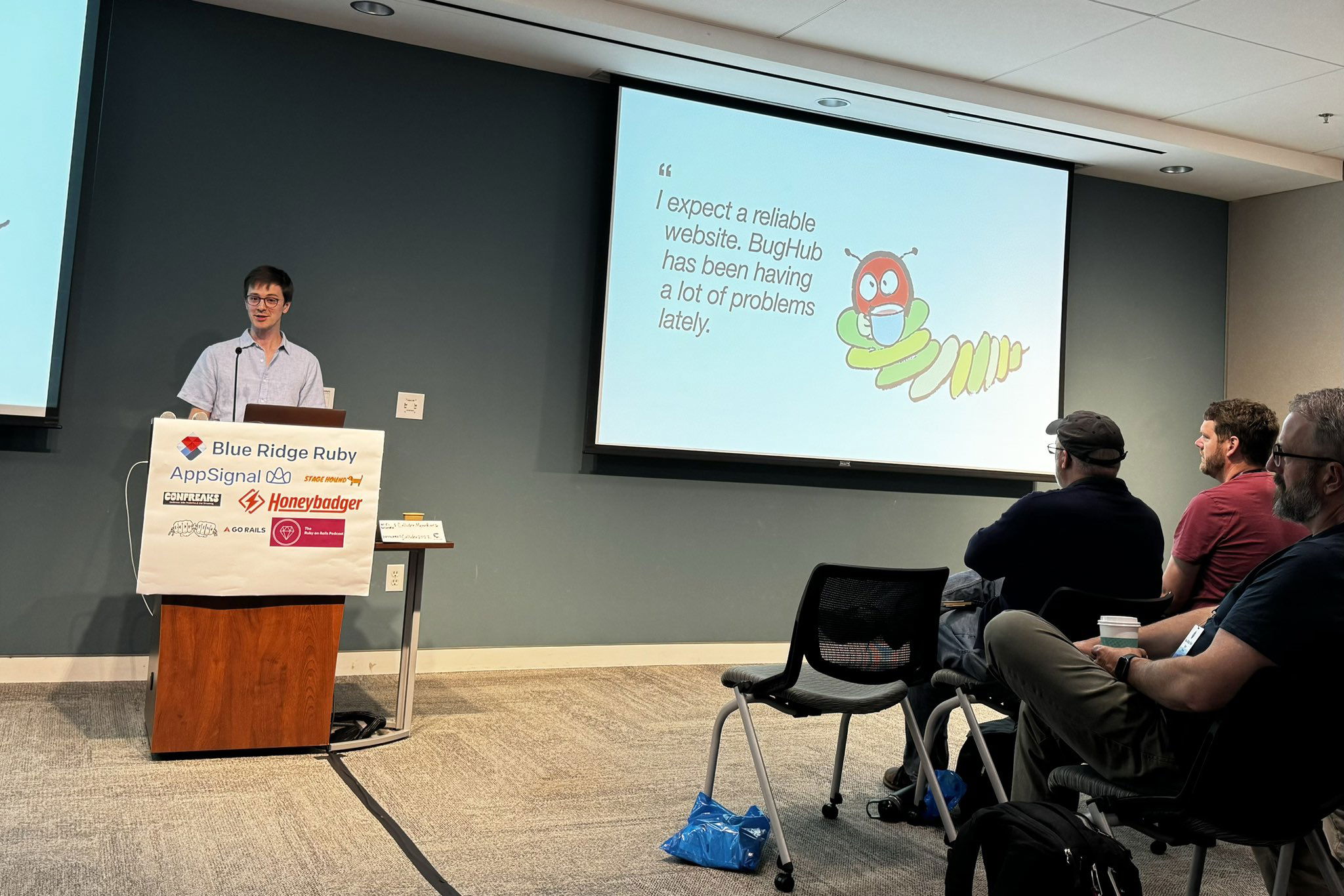 Speaker at a technology conference presenting a slide titled “I expect a reliable website. BugHub has been having a lot of problems lately.” with a cartoon bug image, in front of an audience of attentive attendees.