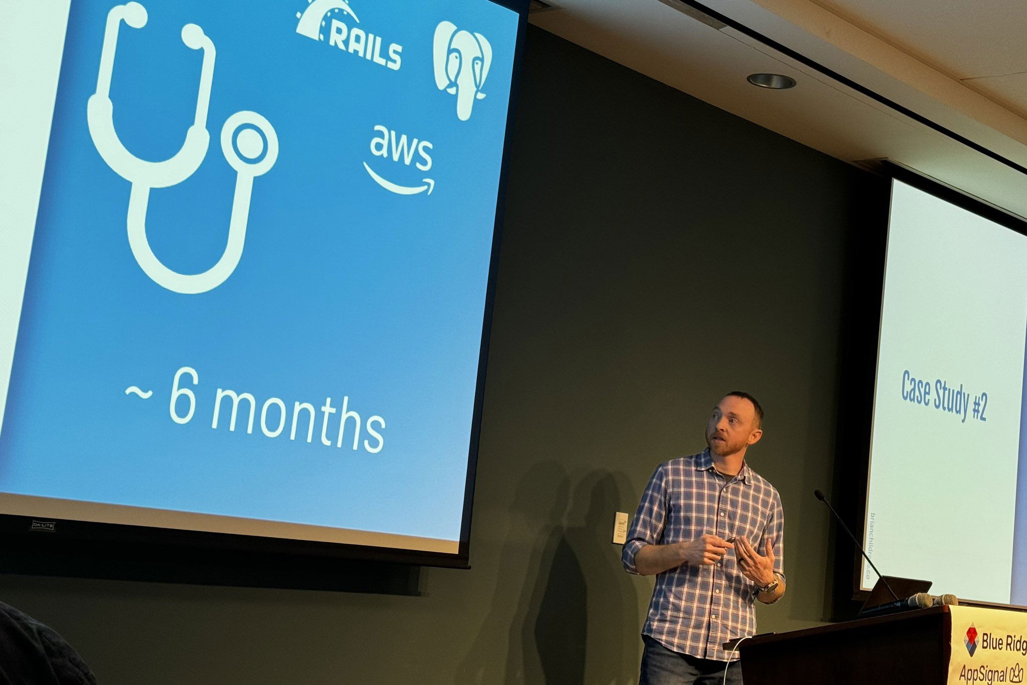 Presenter at a conference discussing a case study related to Rails and AWS, indicated by icons on a slide that reads “~ 6 months”, standing beside a podium with a Blue Ridge Ruby conference banner.