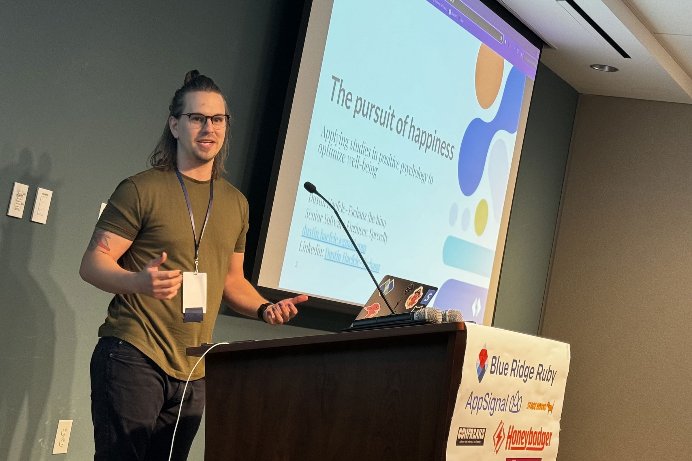 Dustin Haefele-Tschanz presents “The Pursuit of Happiness: Applying Studies in Positive Psychology to Optimize Well-being” at Blue Ridge Ruby conference, engaging the audience with a slide displayed behind him, featuring his contact information and professional title.
