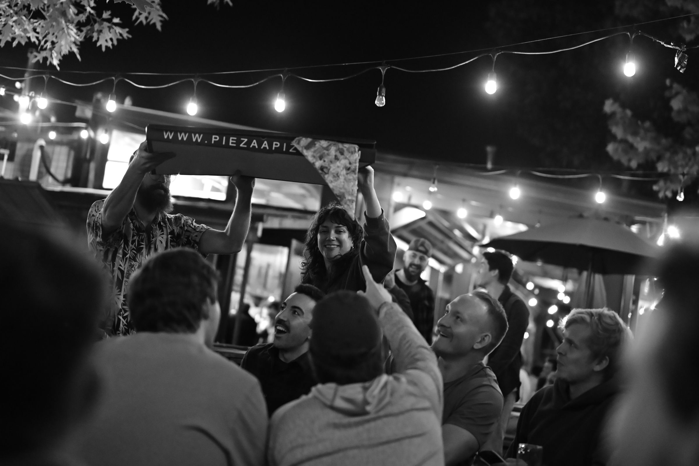 A black and white photo captures a lively scene at an outdoor gathering under string lights. A person in the center is holding up a large pizza box from “Piezaa Pizza” with a slice of pizza in hand, while others around them look up, smiling and pointing at the pizza. The ambiance suggests a casual, festive atmosphere with people enjoying food and company.