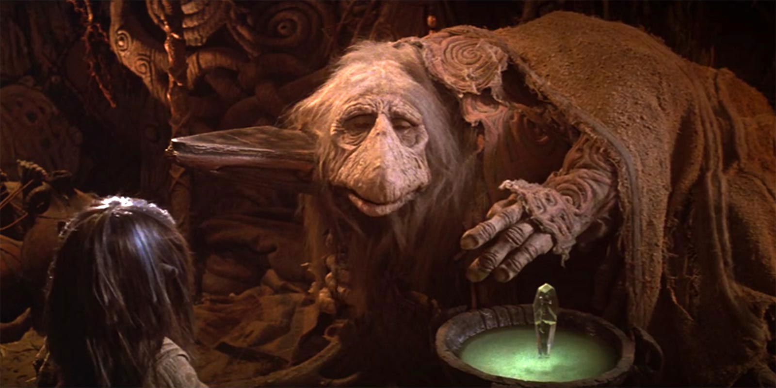 A large, wise-looking creature with long hair and a cloak leans over a glowing crystal in a dimly lit, intricate cave setting, while a smaller figure observes from the foreground.