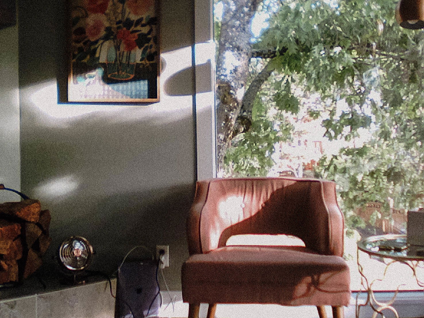 A cozy corner of a room with a pink chair, a framed floral painting on the wall, logs of firewood, and a view of green trees through the window with sunlight streaming in.