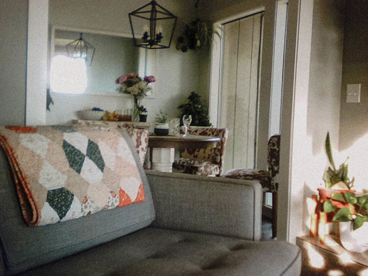 A cozy living room with a gray couch and a colorful quilt, facing a dining area with flowers on the table and pendant lights, and a glimpse of sunlight shining on plants near the doorway.