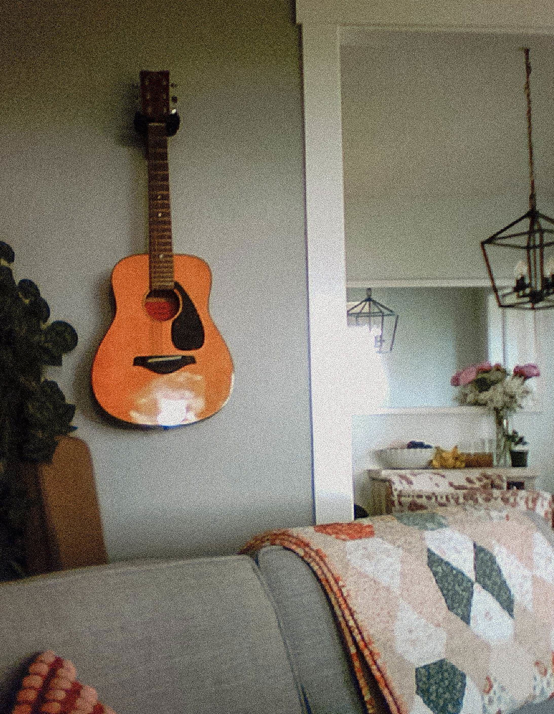 An acoustic guitar hangs on a wall above a gray couch with a quilt, while a dining area with flowers and pendant lights is visible in the background.