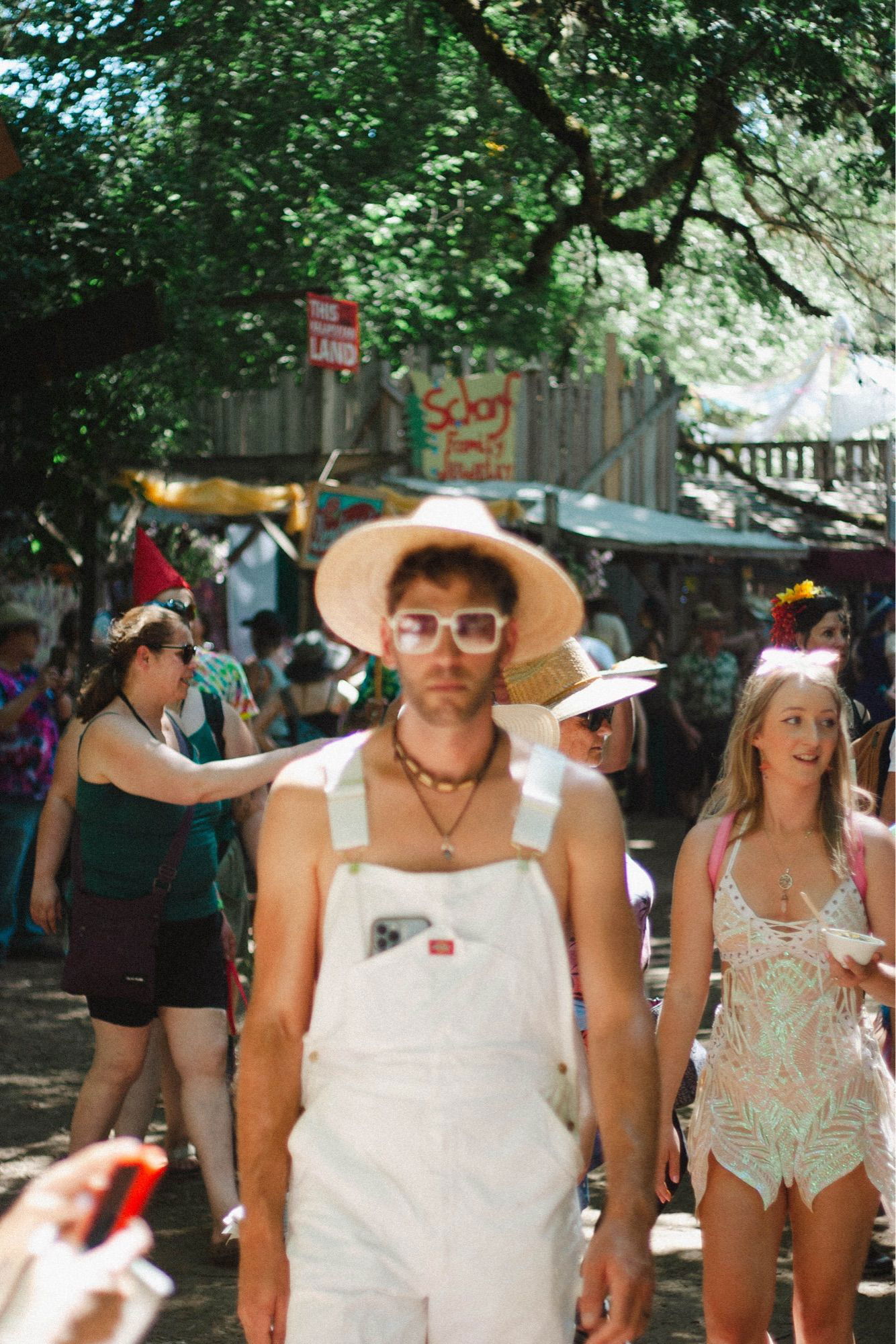 A person at a festival wearing white overalls, a wide-brimmed straw hat, and white sunglasses. They stand amidst a bustling crowd with colorful decorations and signs in the background, including one that reads “This Land” and another for a school family activity.