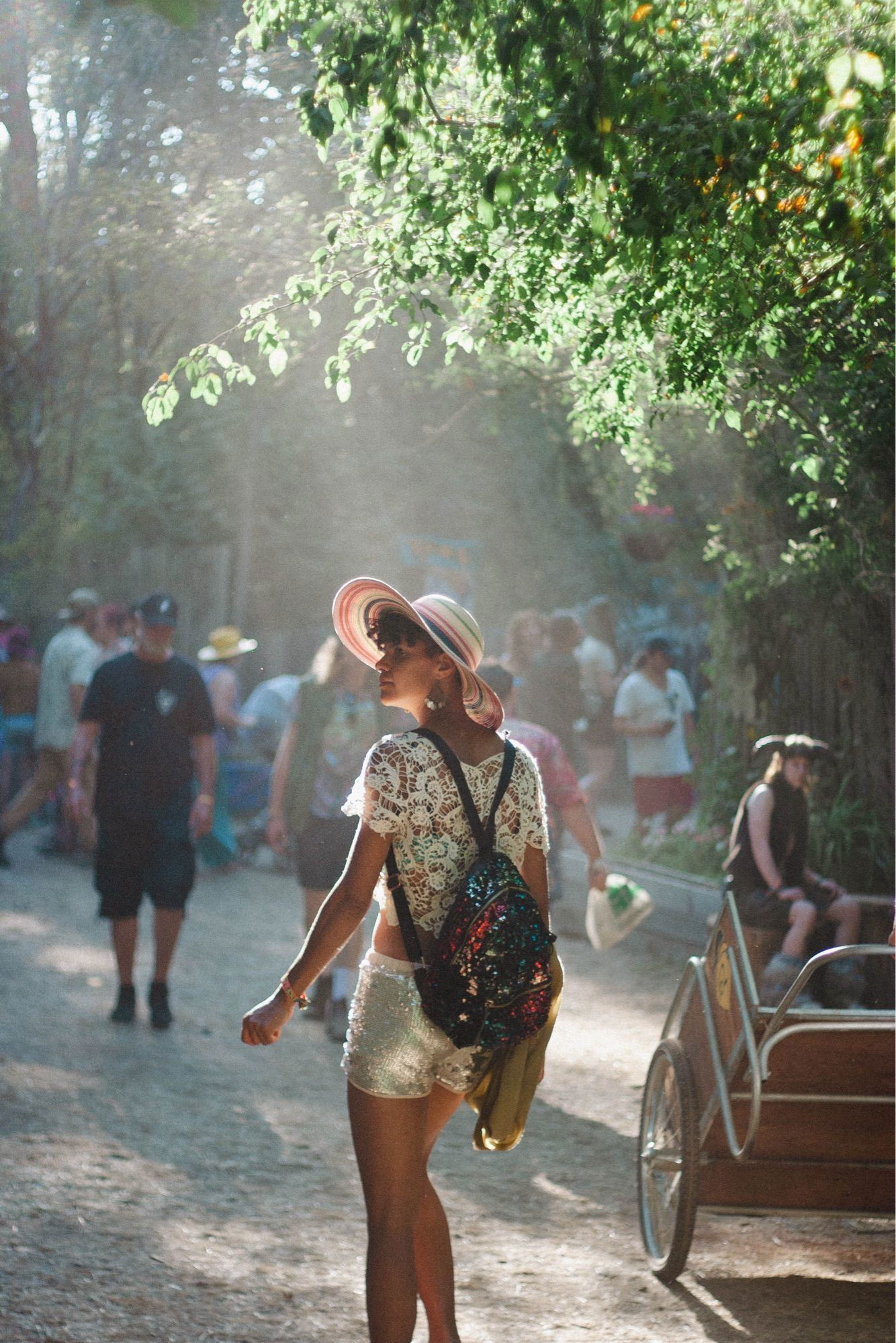 A person walking through a sunny, tree-lined path at a festival. They wear a large pink hat, a white lace top, and shiny silver shorts, with a colorful backpack. The path is filled with other festival-goers, and the sunlight creates a soft, hazy glow through the trees.