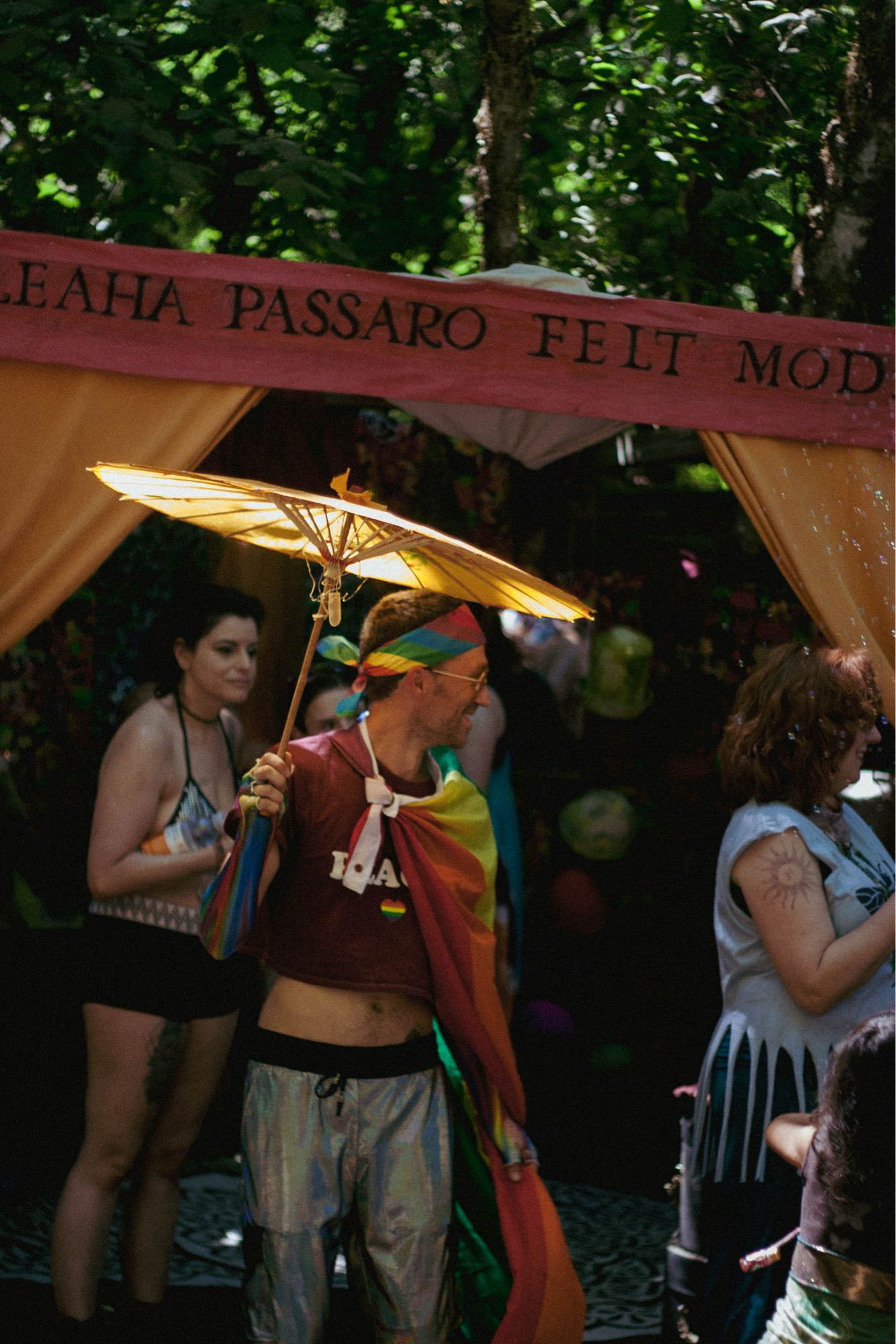 A festival scene with a person holding a yellow parasol and wearing a colorful rainbow outfit. The background includes a booth with a sign reading “Leaha Passaro Felt Mod” and various people enjoying the event.