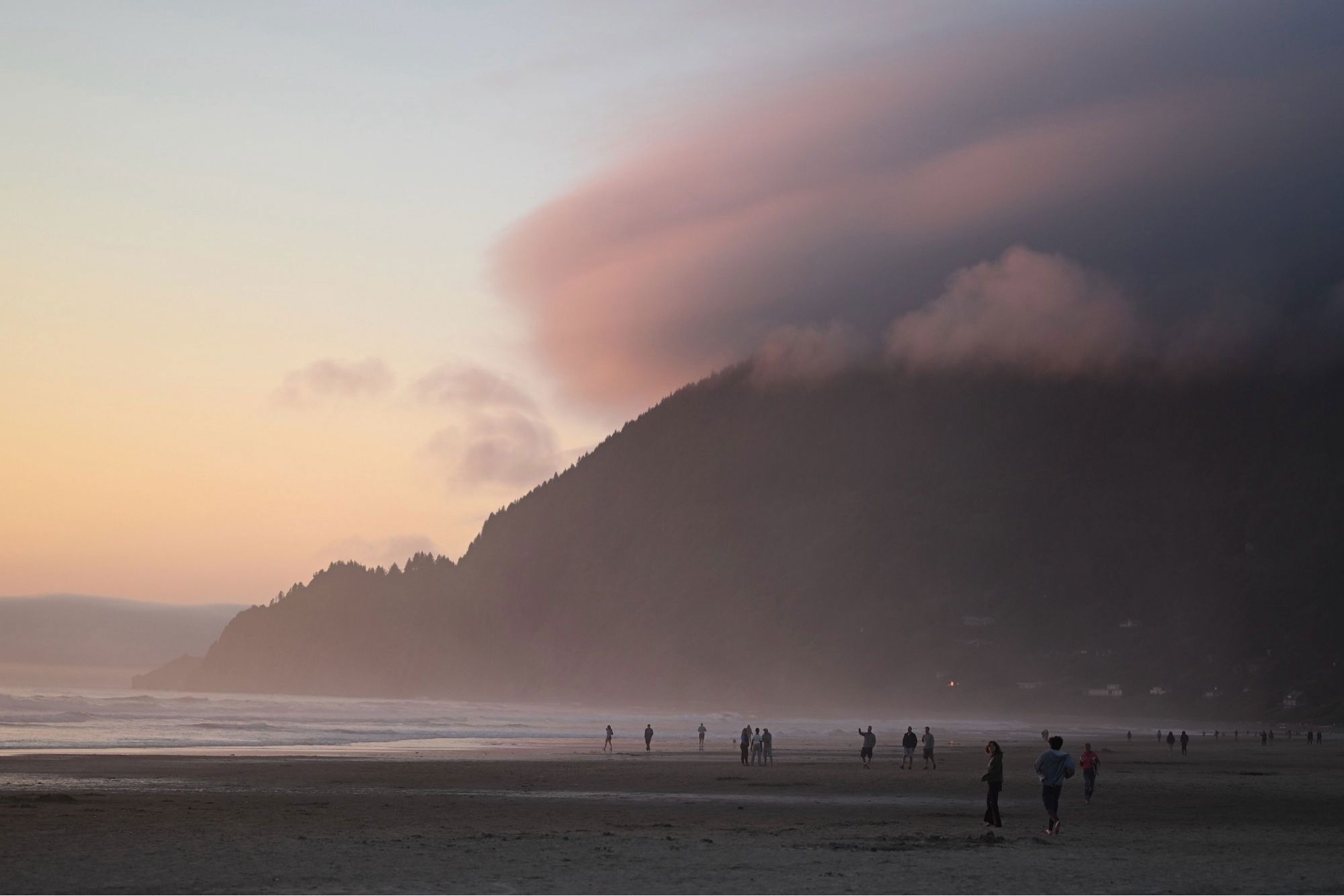 Evening beach scene with a misty mountain in the background, people walking on the sand, and soft twilight colors in the sky.