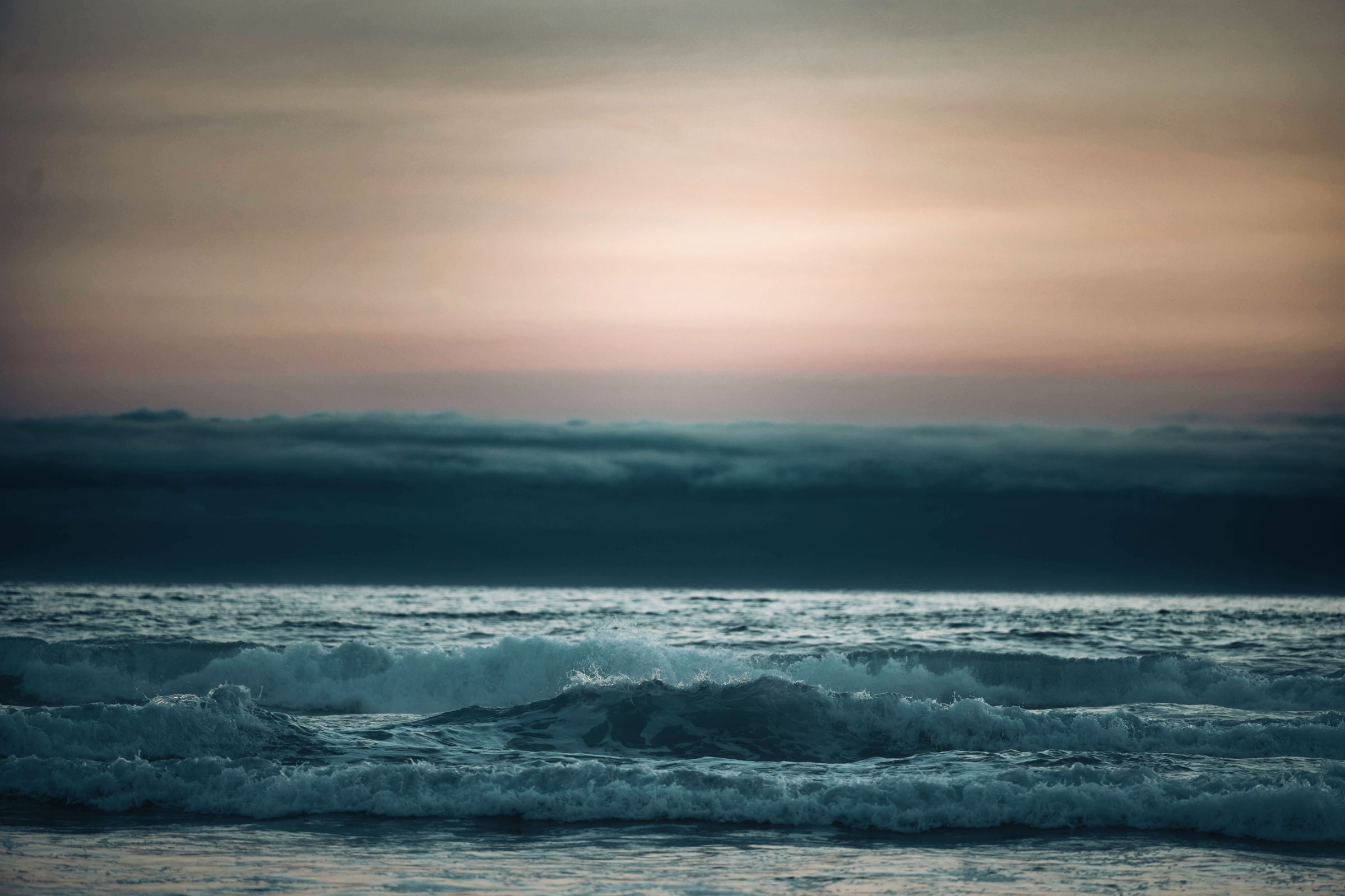 An ocean scene at twilight with gentle waves rolling in under a sky transitioning from warm pink and orange tones near the horizon to cool blue and gray hues, with a layer of low-lying clouds on the horizon—almost like a tidal wave approaching.