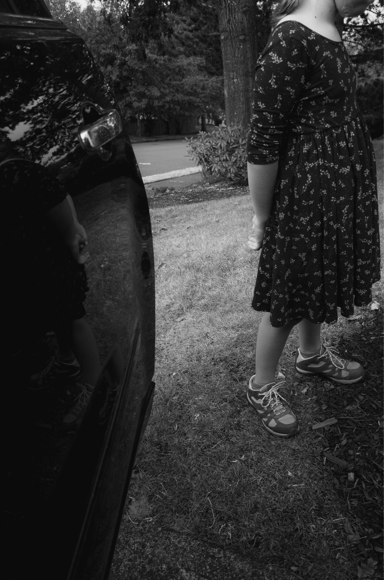 A black-and-white photo shows a young girl in a patterned dress standing partially out of frame, her upper body obscured. She stands on a grassy area next to a parked car, with the reflection of her arm and part of her dress visible in the car's surface. The scene is outdoors with trees and shrubs in the background.