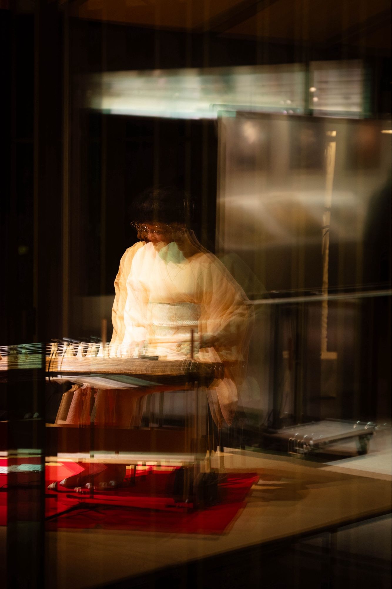 A blurred, ethereal view of a person in traditional Japanese attire playing a koto (a long Japanese zither) through a reflective surface. The image has a warm, golden glow, with the musician’s form softly illuminated against a darker background.