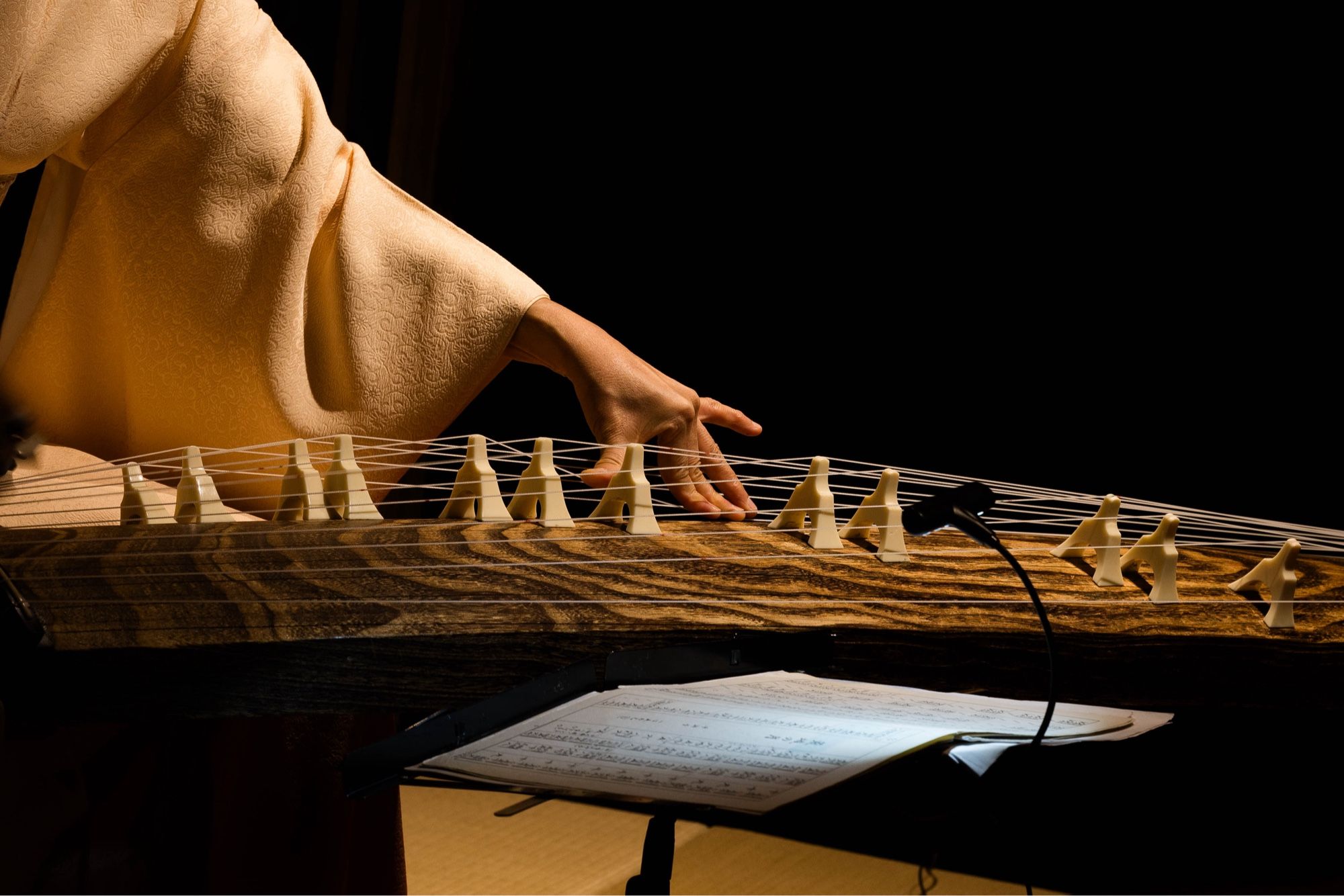 A close-up view of a person’s hand and arm in a cream-colored kimono sleeve, plucking the strings of a koto. The wooden body of the instrument is visible, showing its intricate grain. A sheet of music is partially visible in the foreground.