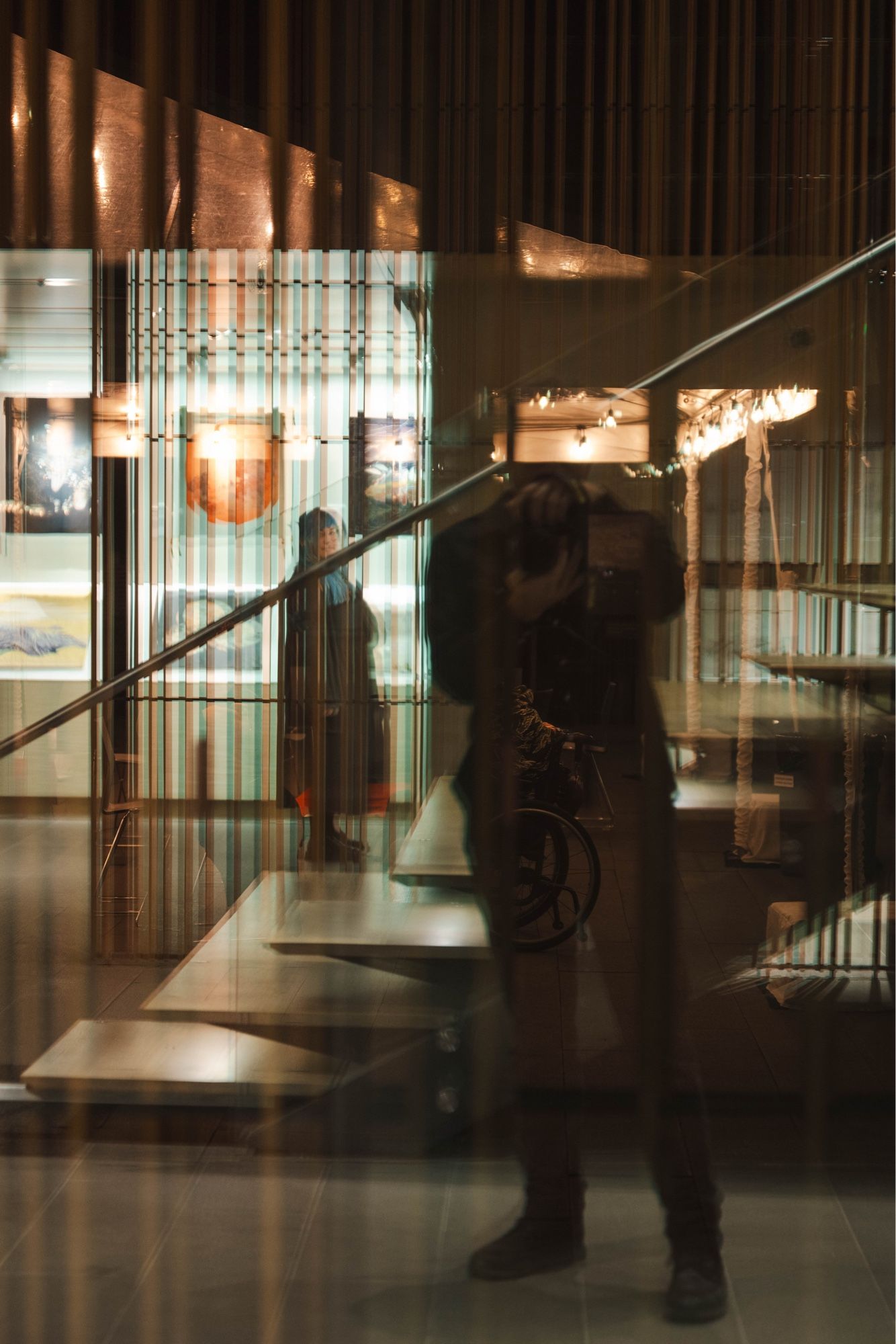 An interior view of a modern building with glass walls and copper-colored ceiling elements. Reflections and lighting create a complex, layered visual effect. A silhouette of a person is visible in the foreground, with displays and lighting fixtures illuminating a second person in the background.