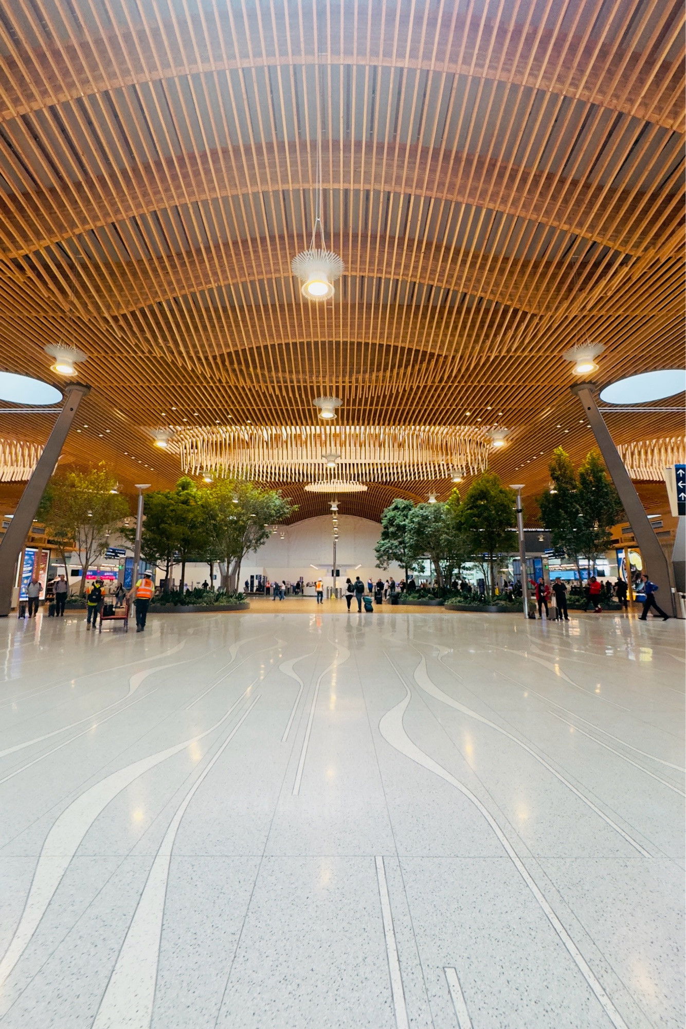 A spacious airport terminal with a curved wooden ceiling, modern lighting fixtures, and indoor trees. People are walking on the reflective floor, and signage is visible in the background.