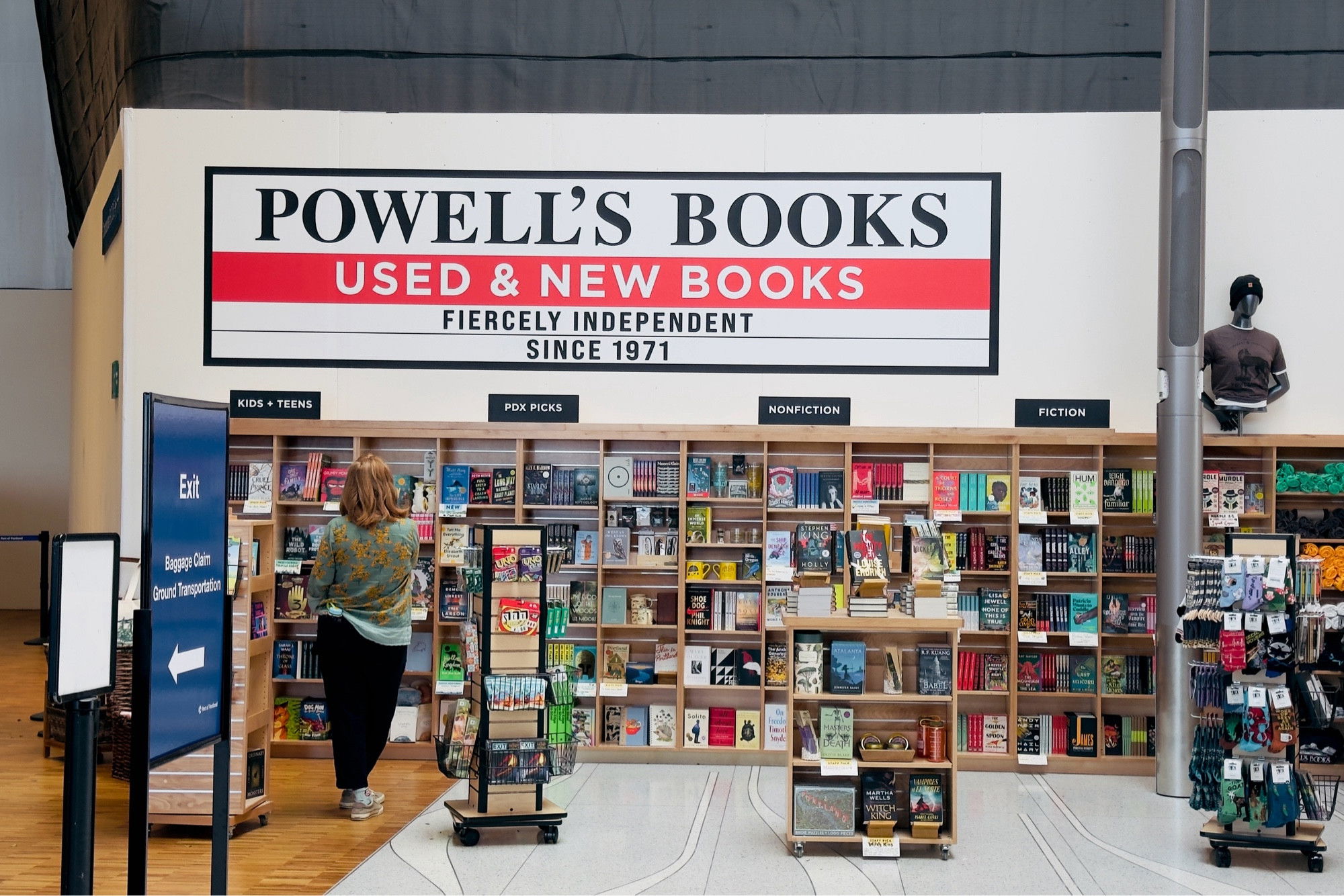 A Powell’s Books store in an airport. The storefront has a large sign reading “POWELL’S BOOKS - USED & NEW BOOKS - FIERCELY INDEPENDENT SINCE 1971”. Bookshelves filled with colorful books are visible, and a person is browsing the selection.