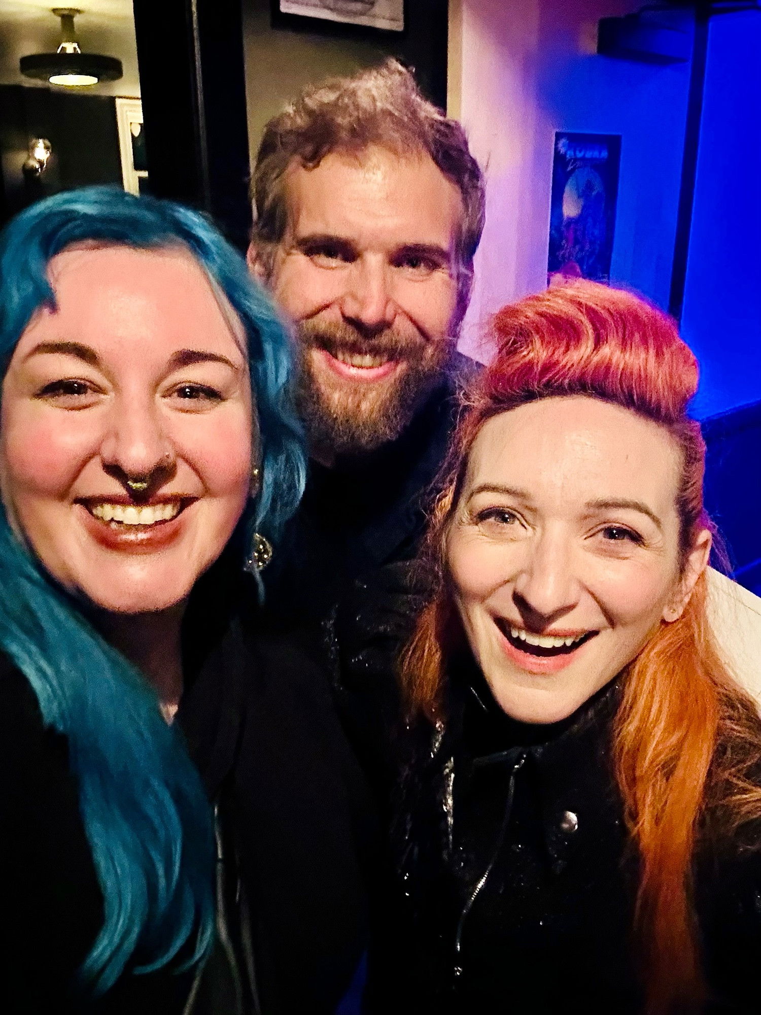 Three smiling people pose for a selfie. On the left is a woman with blue hair, in the middle is a bearded man, and on the right is Shara Nova with orange hair. The background shows the colorful lighting of a venue interior.​​​​​​​​​​​​​​​​