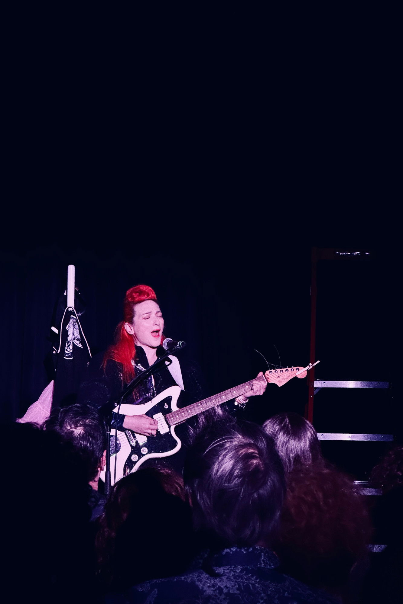 A musician with bright orange hair plays an electric guitar and sings into a microphone on stage. She wears a dark outfit and is bathed in pink light, with the crowd visible in the foreground.