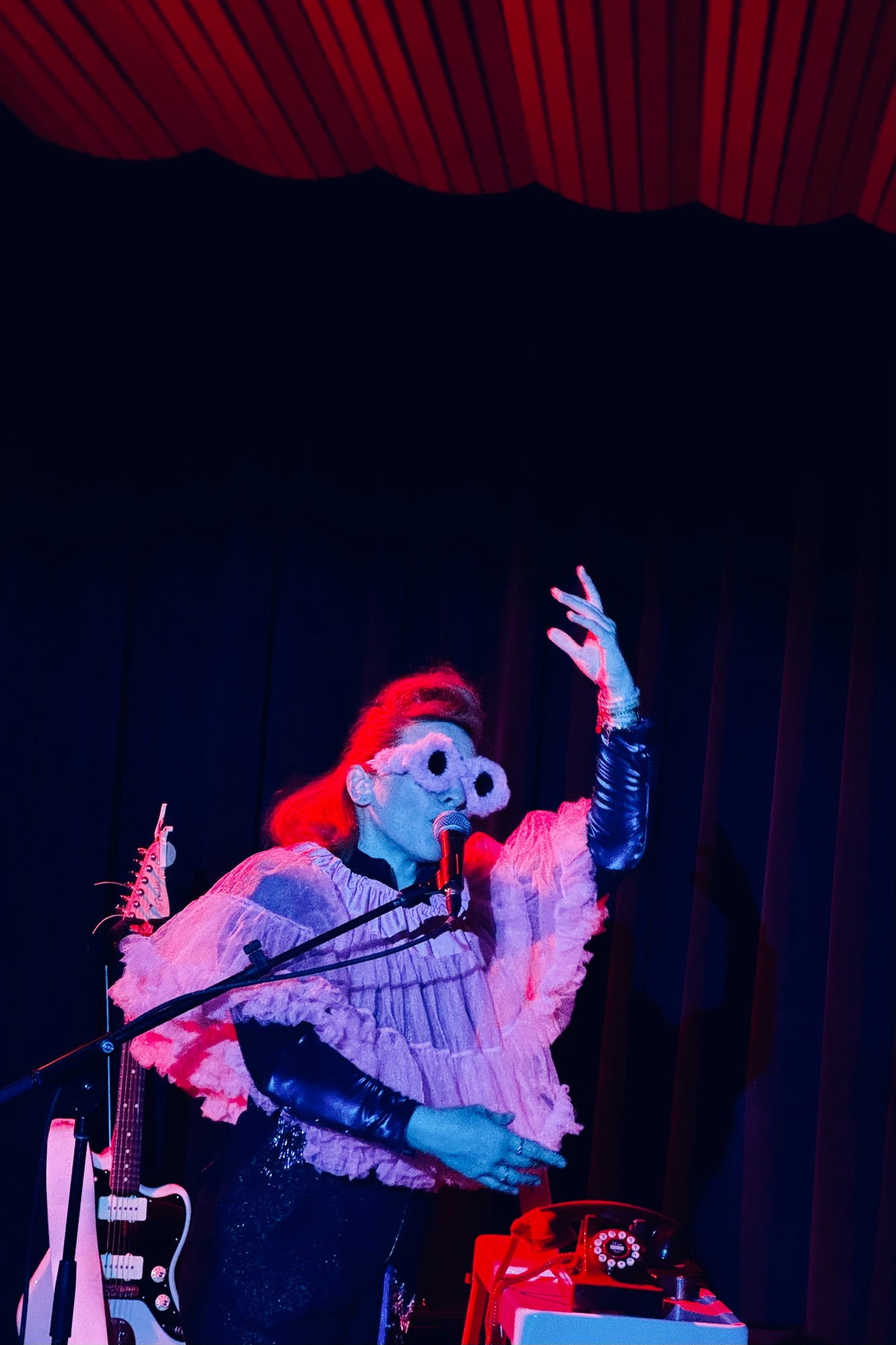 A performer in a ruffled pink top, sun glasses, and dark pants sings into a microphone on stage with their hand raised dramatically. The stage is lit with red and blue lights, with a red curtain visible above.