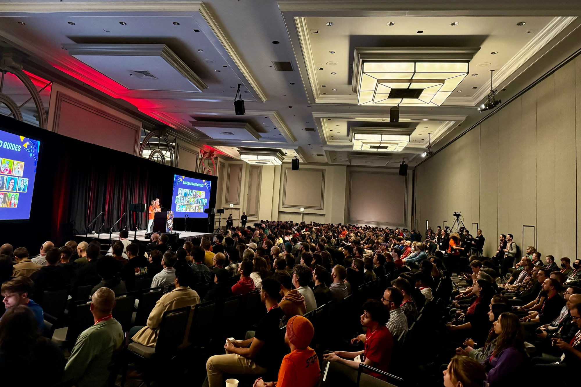 A large conference or presentation room filled with rows of attendees watching a stage presentation. The room features an elegant coffered ceiling with recessed lighting and architectural details. At the front, there are two large projection screens displaying what appears to be gaming or streaming content with colorful character portraits. The stage is elevated and has a black curtain backdrop with red accent lighting. The audience, sitting in theater-style seating, appears to be predominantly young adults, and the room is at full capacity. Professional lighting and camera equipment can be seen around the edges of the space, suggesting this is a major event or convention.​​​​​​​​​​​​​​​​