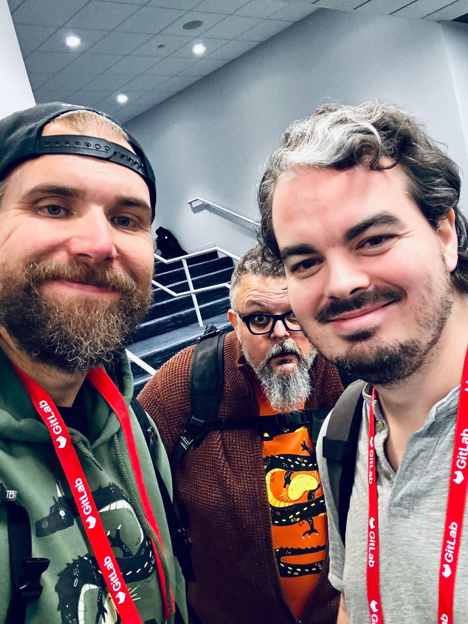 Three men in casual attire posing for a selfie in a conference setting, all wearing conference lanyards: one in a backwards black cap and green hoodie, another in glasses and a brown sweater making a playful expression, and a third in a gray shirt, all sporting various styles of facial hair.​​​​​​​​​​​​​​​​