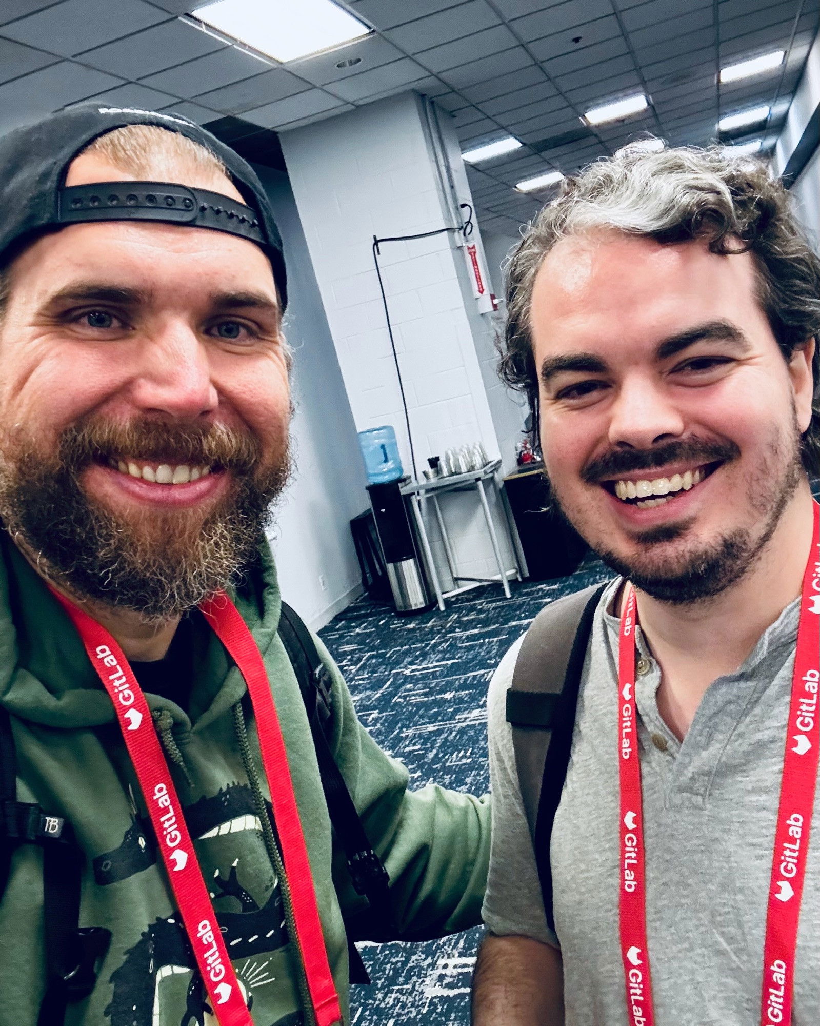 Two men smiling at the camera in a conference space, both wearing conference lanyards - one in a backwards black cap and green hoodie with a full beard, and another in a gray henley shirt with dark wavy hair and facial hair. 