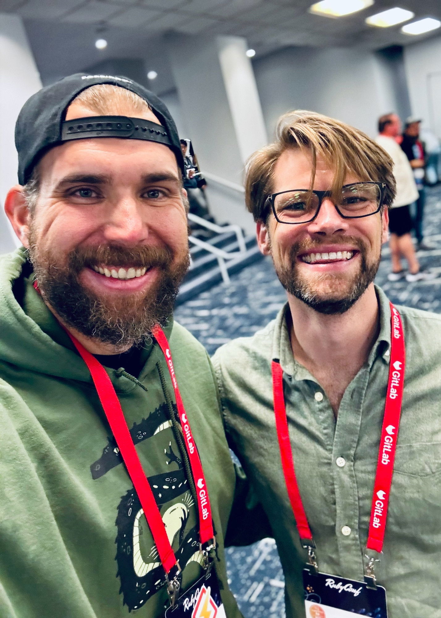 A selfie of two men at RubyConf - one in a backwards black cap and green hoodie with a full beard, and another with styled hair, glasses and a sage green button-up shirt, both wearing red RubyConf lanyards and smiling broadly at the camera with patterned carpet visible in the background.​​​​​​​​​​​​​​​​