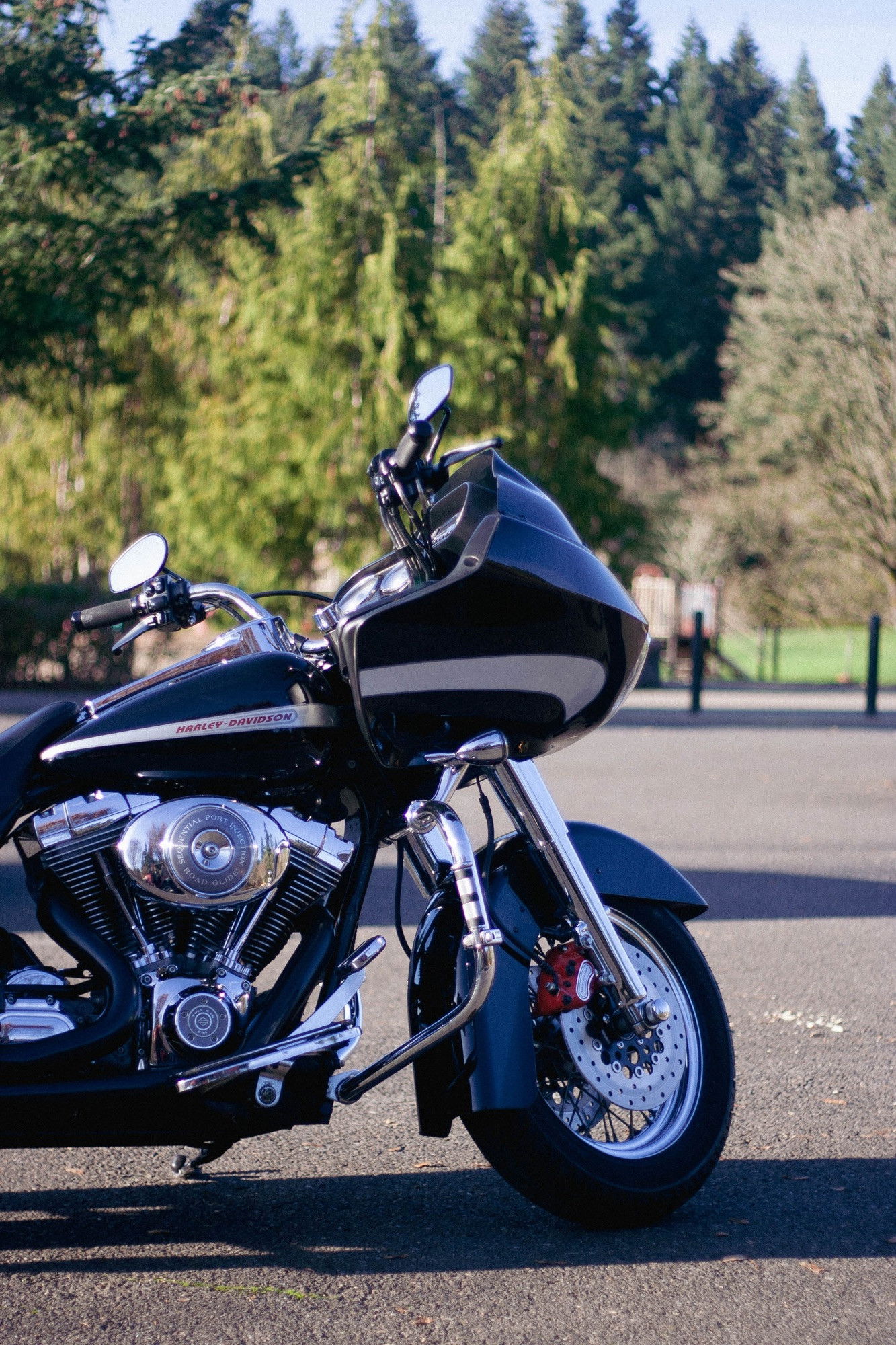 A color photo of a clean black and chrome Harley-Davidson road bike with a creme colored stripe parked in in a parking lot with green trees in the background. 