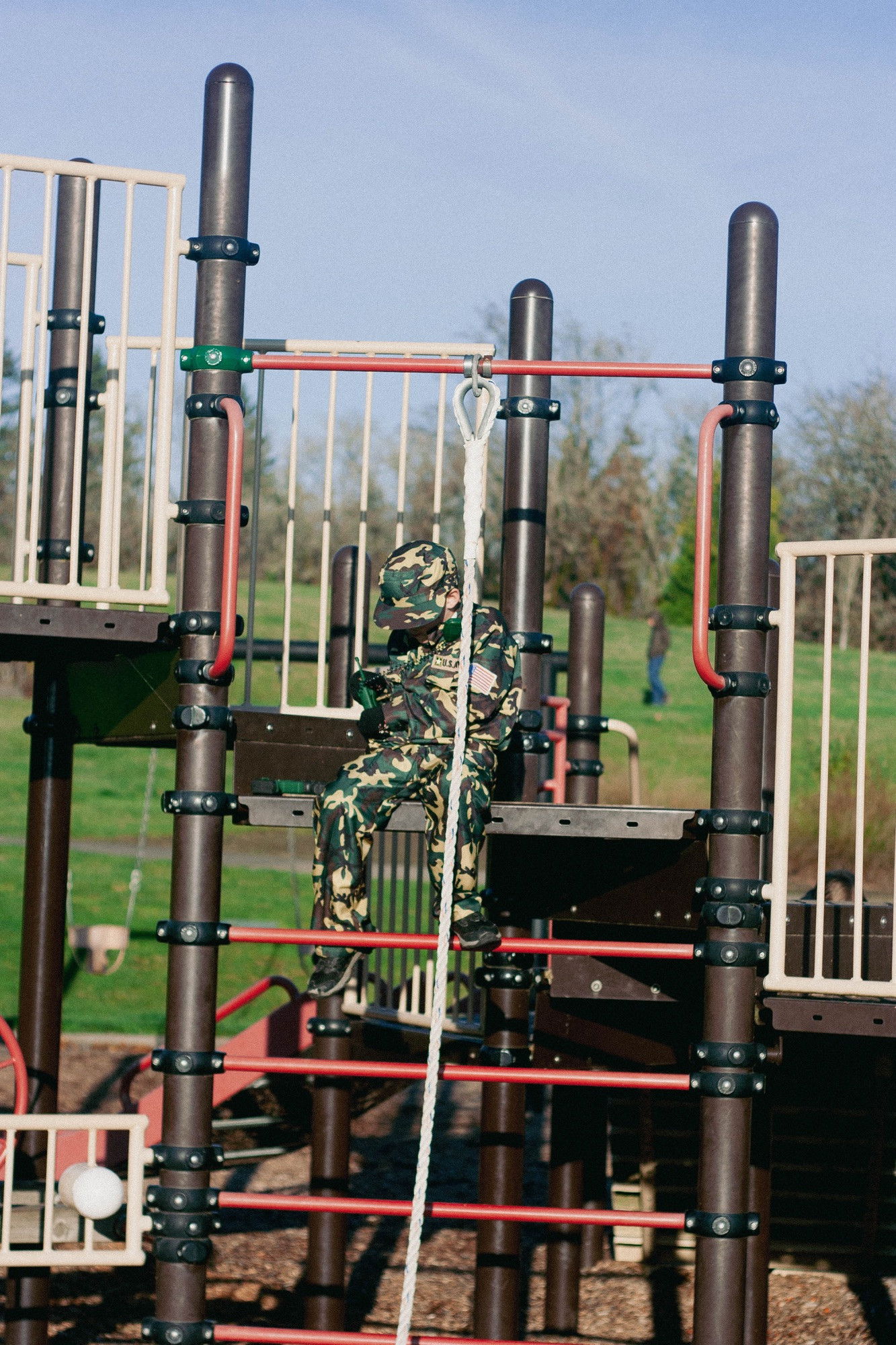 A child in full camouflage military-style clothing with an American flag patch sits on a modern playground climbing structure made of brown poles and red bars, with a rope hanging nearby.