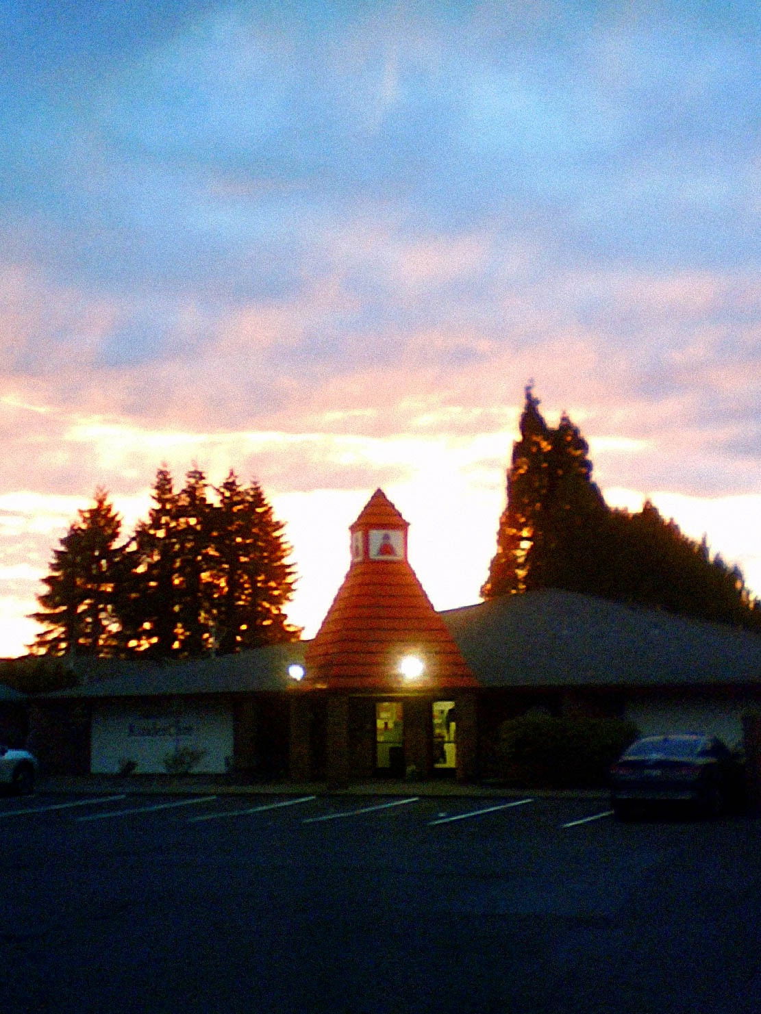 A single-story commercial building with a distinctive red pyramid-shaped cupola or tower on its roof, photographed at dusk. The building has illuminated windows and is framed by tall evergreen trees against a sunset sky with pink and blue clouds. A parking lot is visible in the foreground.