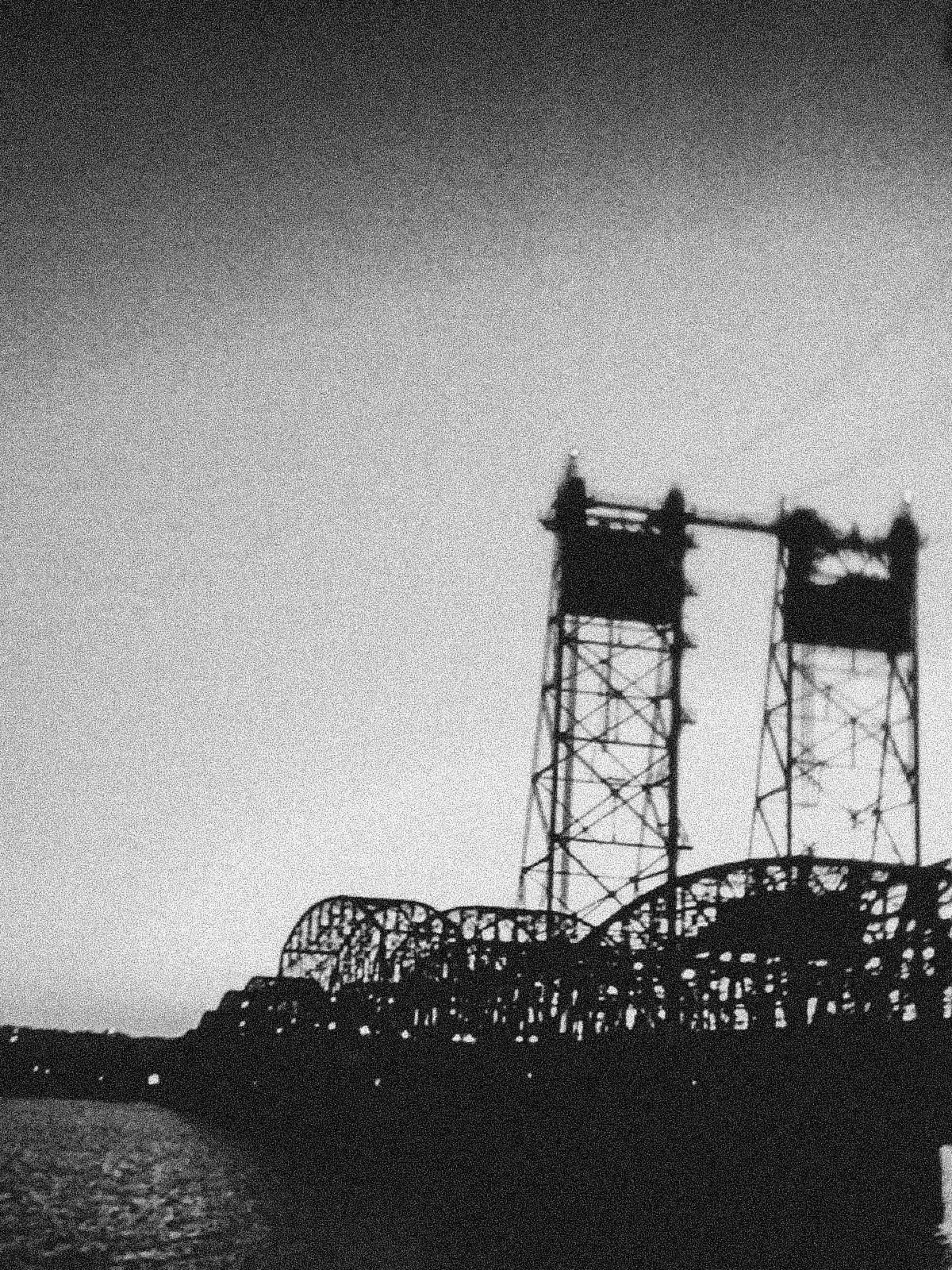 A black and white photograph of a bridge silhouetted against a bright sky, featuring distinctive vertical lift towers and a steel truss structure. The bridge's intricate lattice work creates a complex pattern of shadows and light, with water visible below.