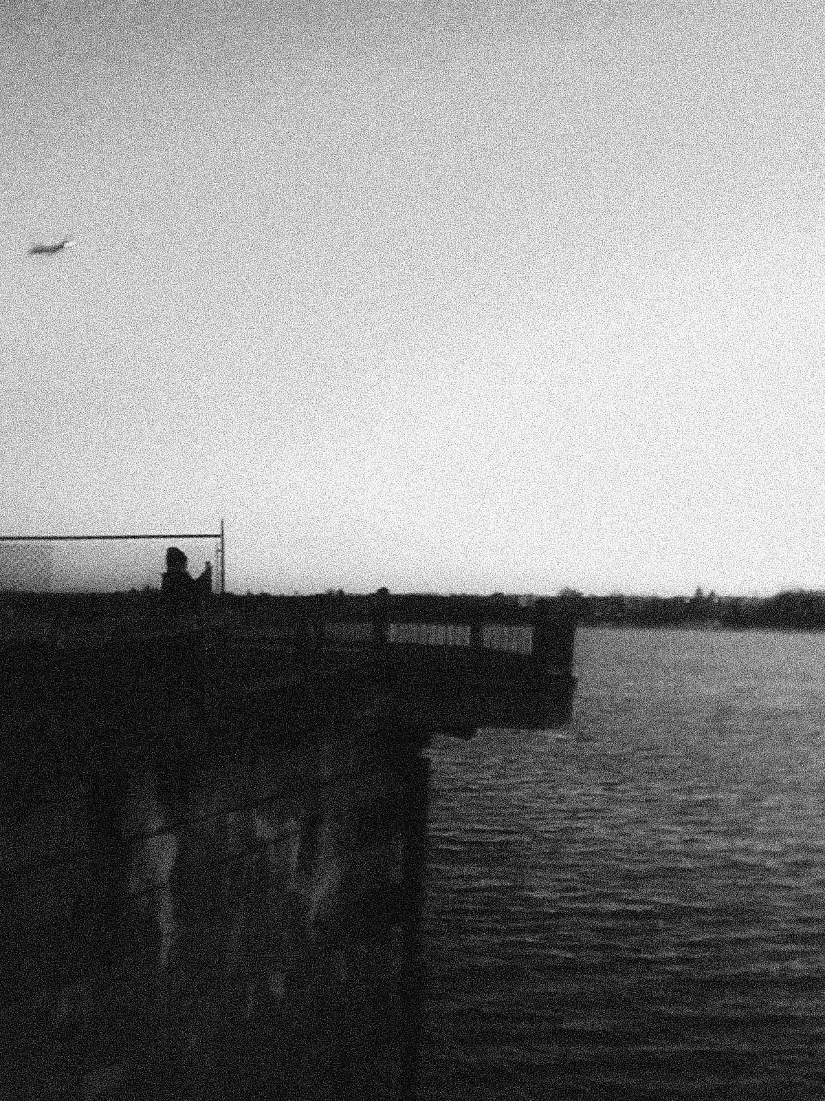 A high-contrast black and white photograph showing a silhouetted figure standing near a chainlink fence along a waterfront, with an airplane visible in the distant sky. The image has a grainy, film-like quality with stark light and dark contrasts.