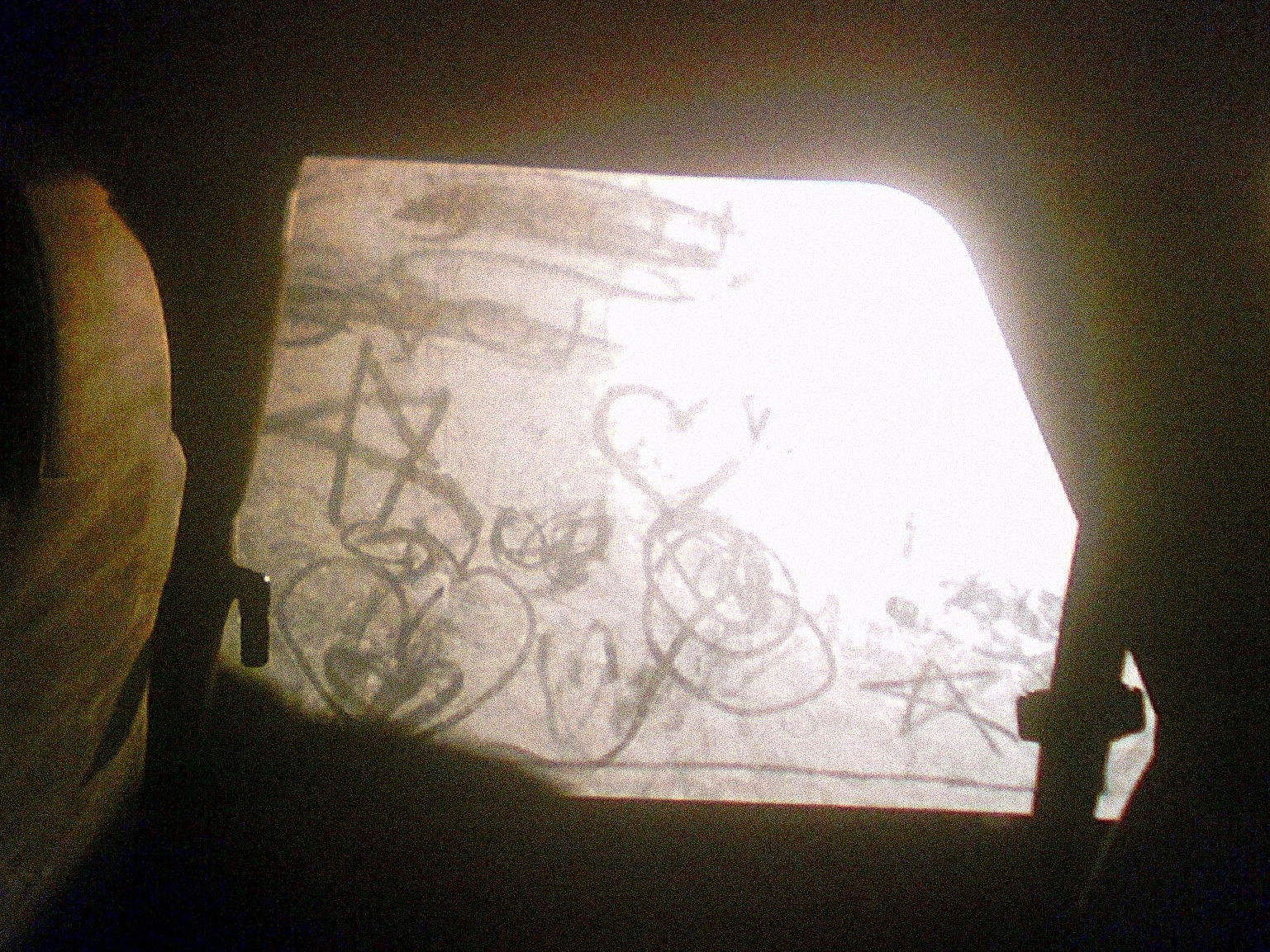 A view through a car window where someone has drawn doodles and marks by finger in the accumulated dirt on the glass, backlit by bright daylight which contrasts the drawings against the dark interior of the car.