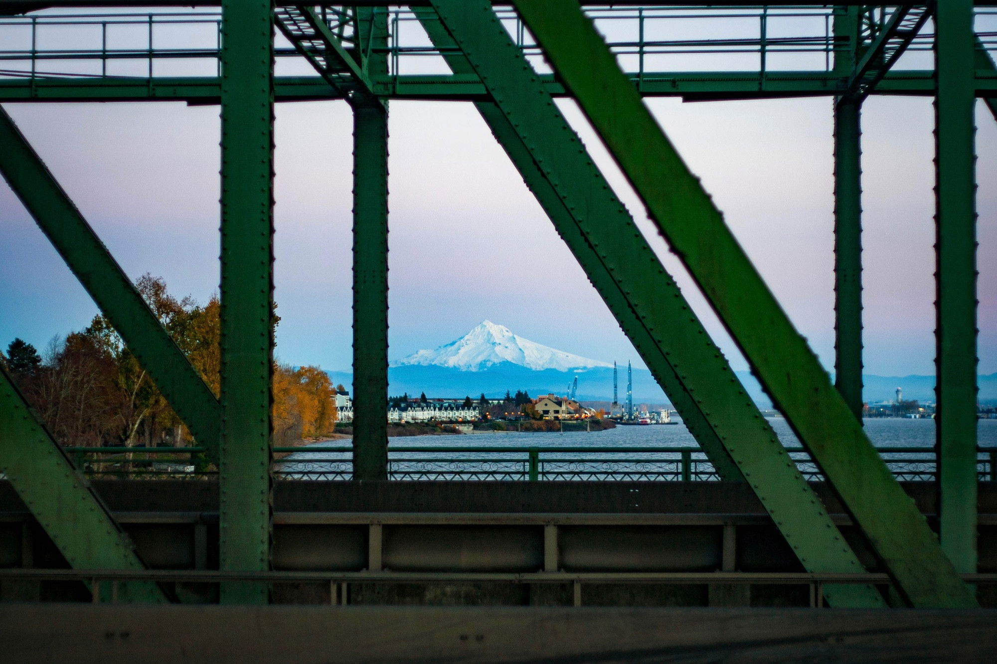 A view of Mount Hood framed by the green steel girders of a bridge, with the Columbia River and industrial waterfront visible in the middle ground, including buildings and crane structures against a crisp winter sky.