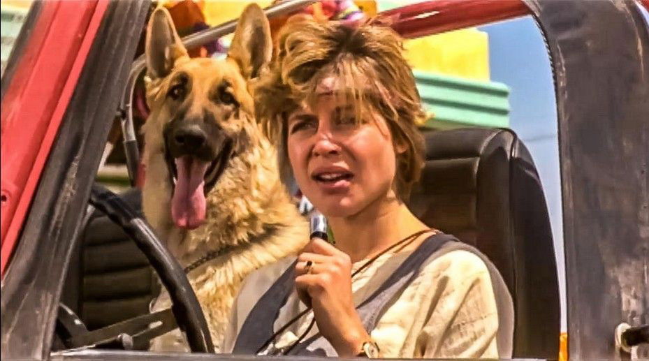 A still from "Terminator" showing Sarah Connor (Linda Hamilton) with messy, windswept hair sitting in a red Jeep Wrangler with a German Shepherd dog as her passenger. Both are viewed through the driver's window, with Sarah appearing youthful and the dog looking alert with its tongue out, against a colorful background.