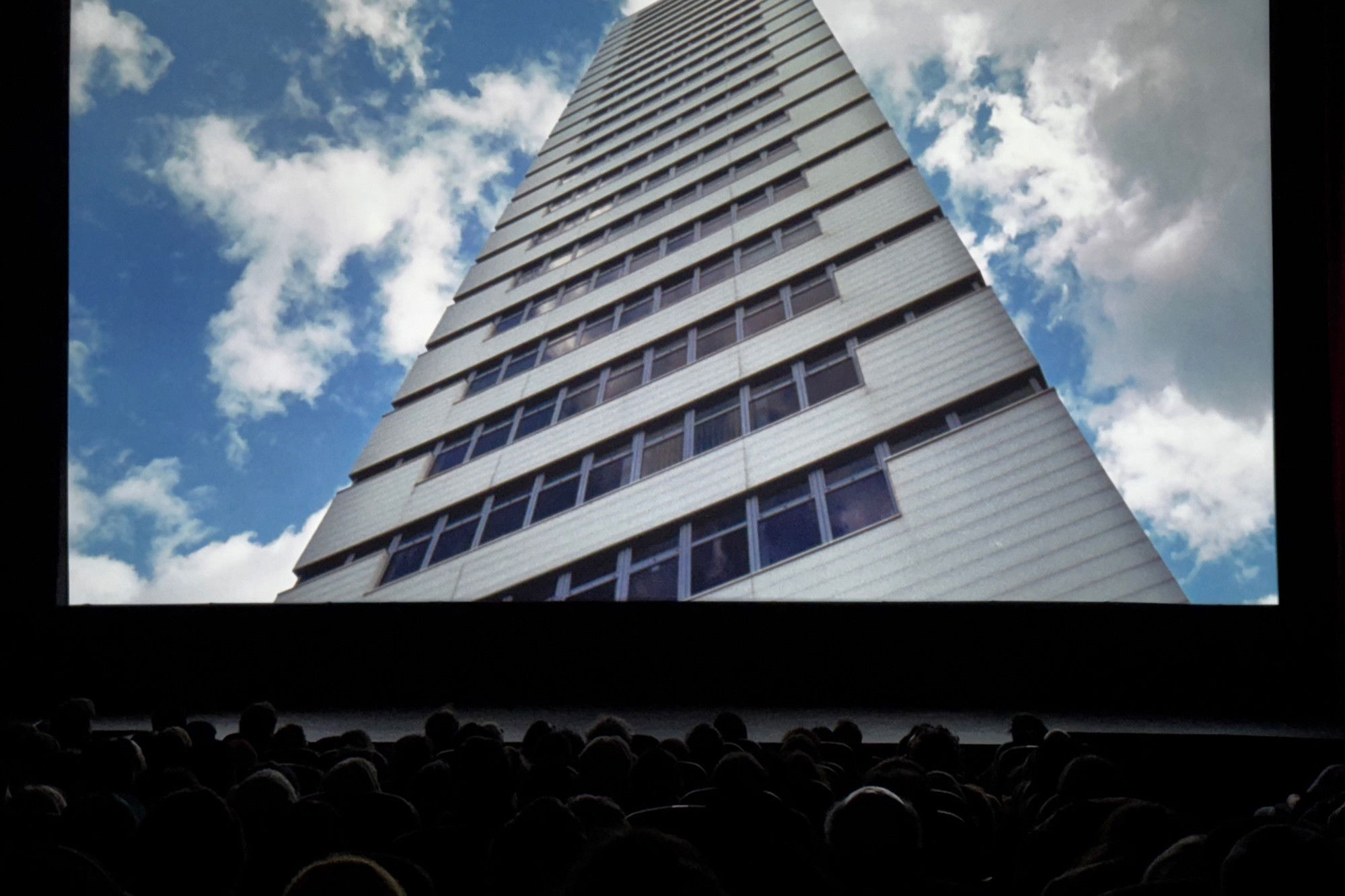 A movie theater audience in silhouette watches a screen showing an upward-angled view of a tall white modernist skyscraper against a blue sky with white clouds.