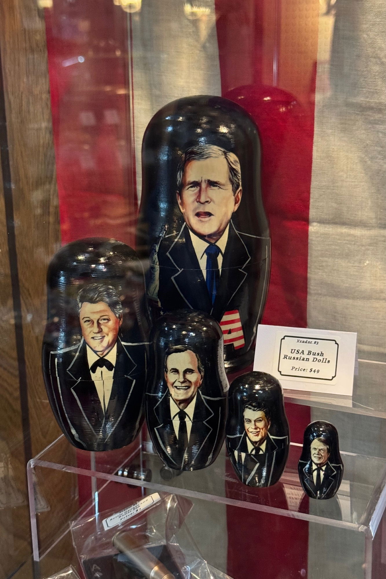 Set of black Russian nesting dolls (matryoshka) displayed in a clear case, featuring painted portraits of U.S. presidents in formal attire. A small display card indicates 'USA Bush Russian Dolls' with a price of $49. The display has red and white backdrop elements.