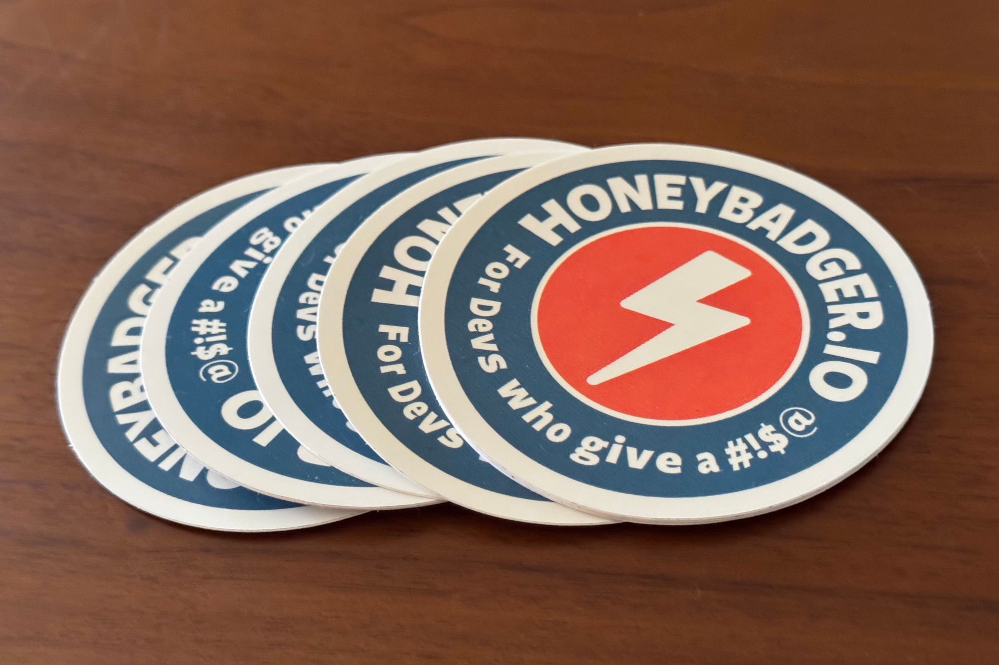 Stack of Honeybadger promotional stickers with the company’s orange and white lightning bolt logo and text reading ‘For Devs who give a #!$@’ on a blue circular background. 