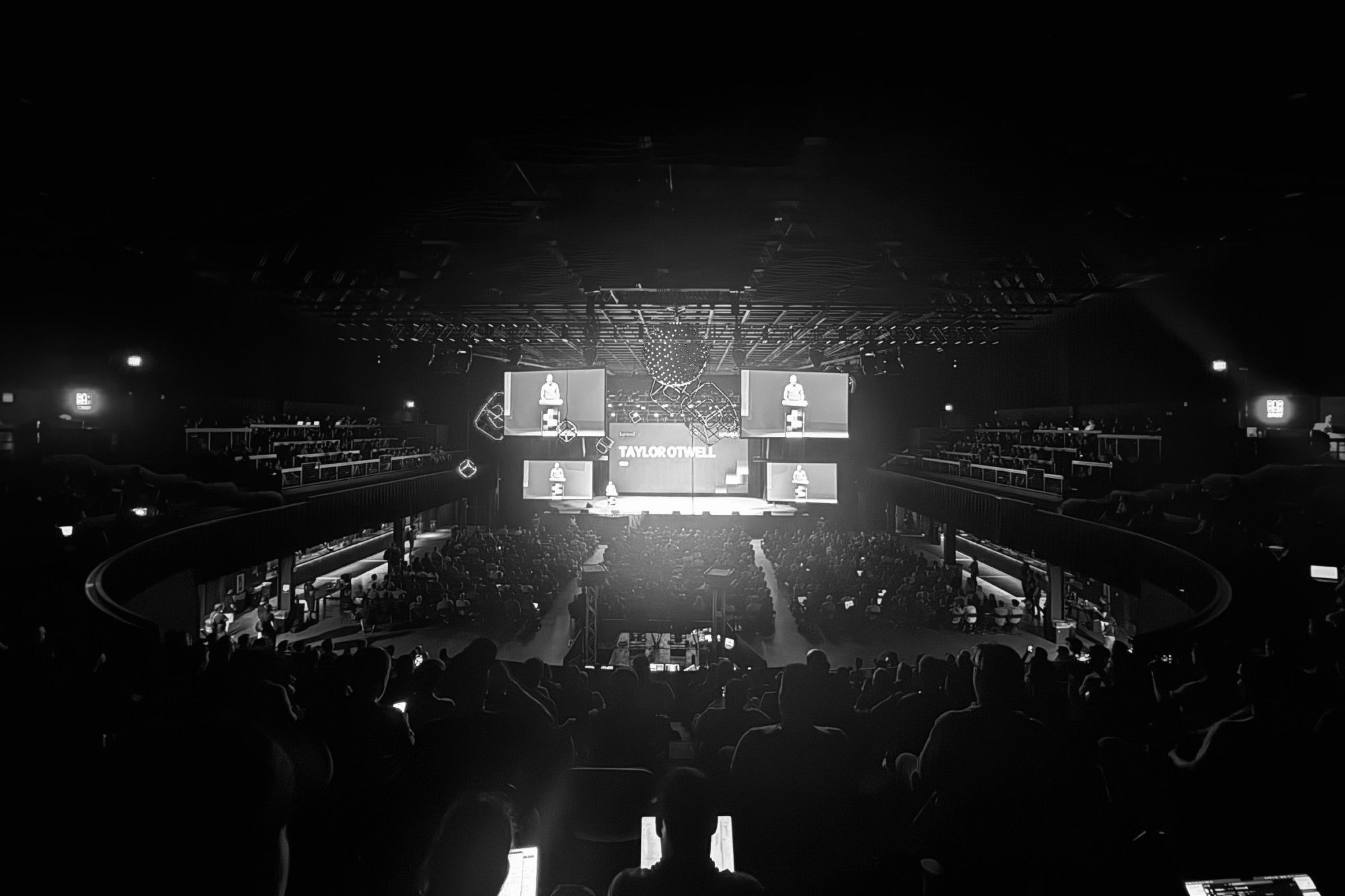 A black and white photograph of a large concert venue filled with audience members, featuring a stage with large video screens displaying "Taylor Otwell" and a man standing at a podium.