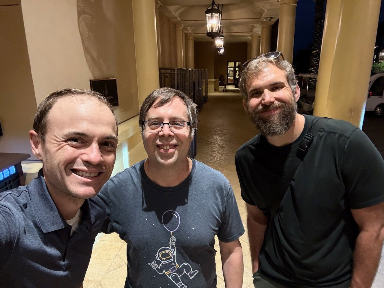 Three men smiling for a selfie in an elegant hotel hallway with columns and hanging lanterns visible in the background. The group includes a man in a polo shirt on the left, a man with glasses wearing a graphic t-shirt in the center, and a bearded man in a black shirt on the right.