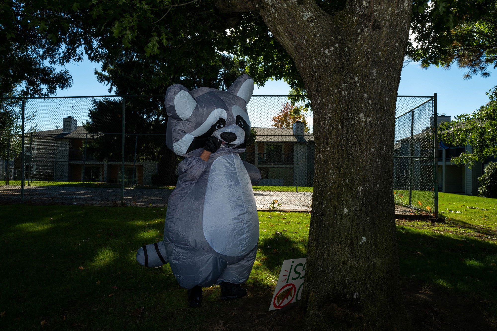A person in an inflatable raccoon suit stands by a tree talking on the phone through the fabric