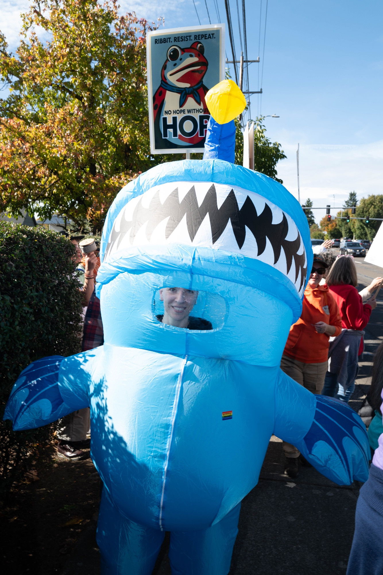 A smiling person inside an inflatable costume. Behind them a sign reads 'RIBBIT. RESIST. REPEAT. NO HOPE WITHOUT HOPE' with a frog illustration