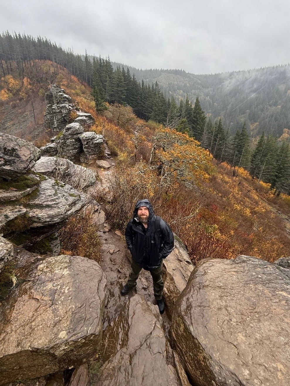 A man standing on a hiking trail with his hands in the pockets of his rain jacket framed by large boulders with yellow shrubs and evergreen trees in the distance