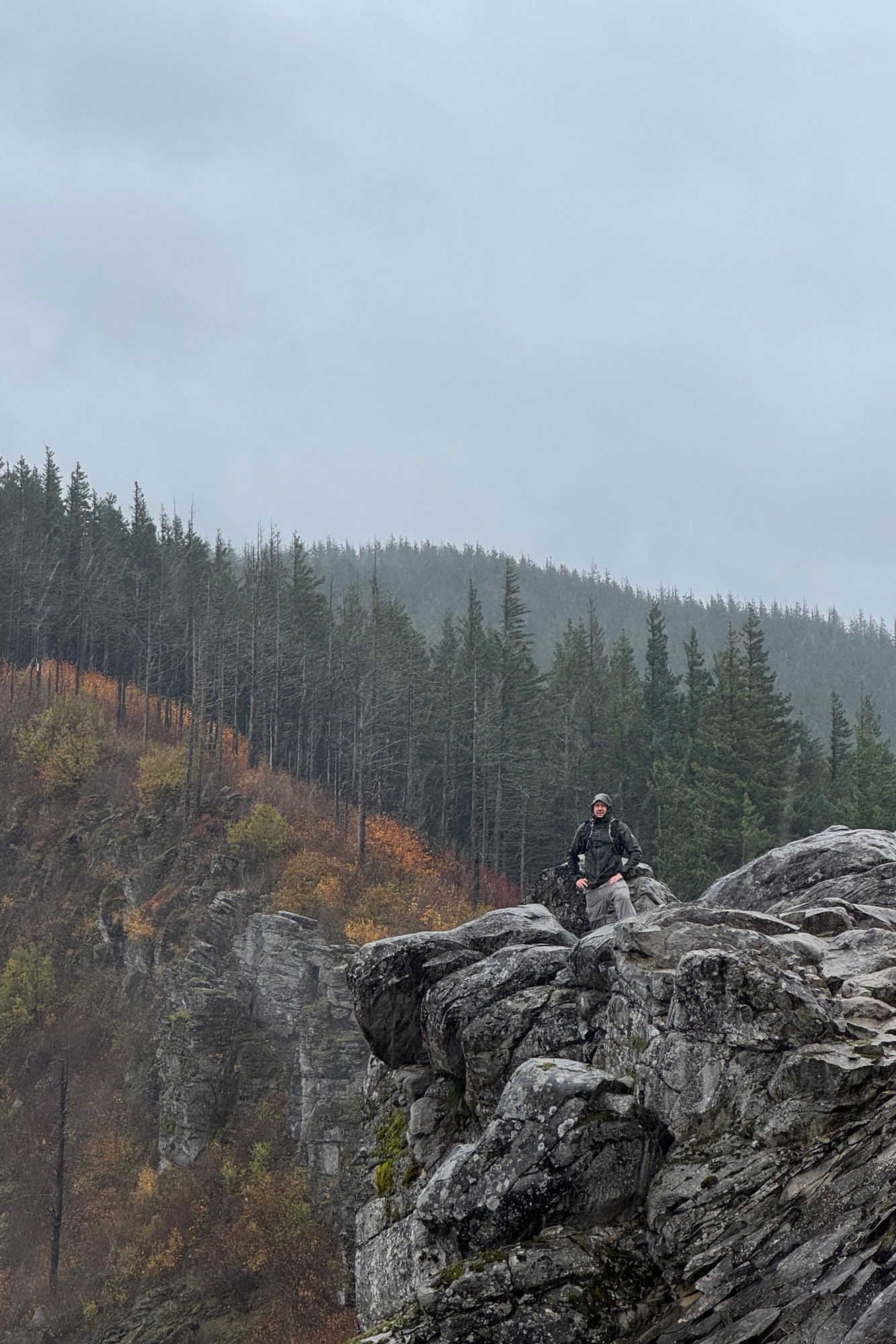 A man standing standing on an outcropping of rock in the distance, with evergreen trees and hills in the in the background