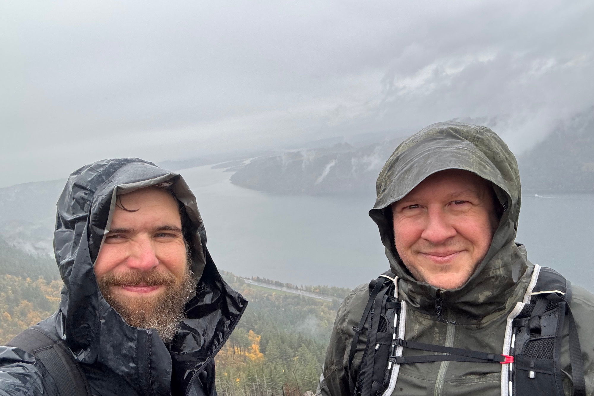 Two men in rain gear smiling for a selfie with the Columbia River in the background