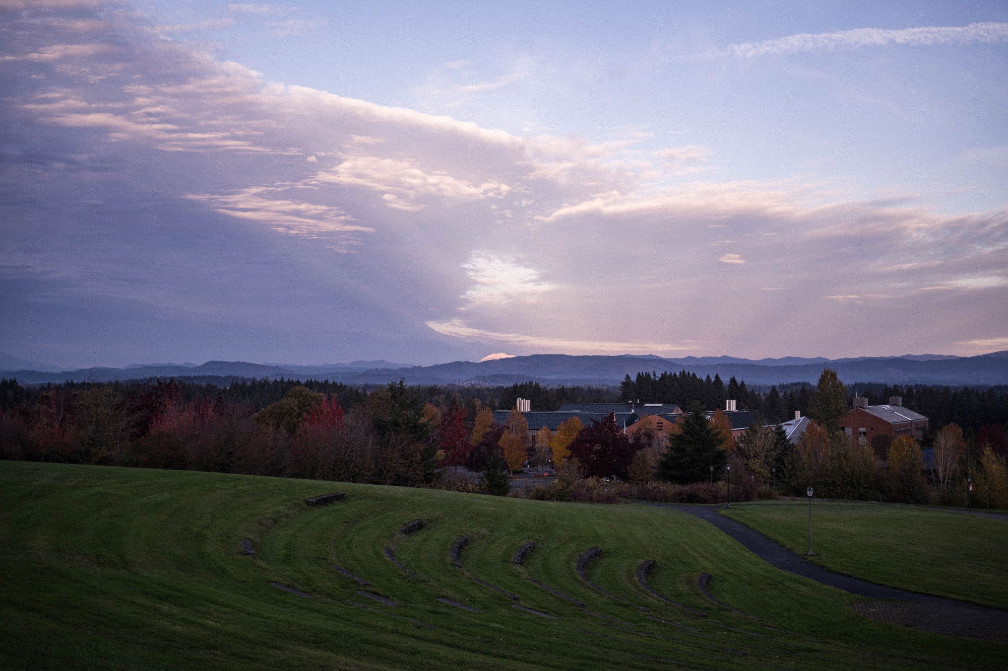 A green hillside with campus buildings and autumn trees in the middle ground with a mountain range under dramatic twilight sky in the distance.