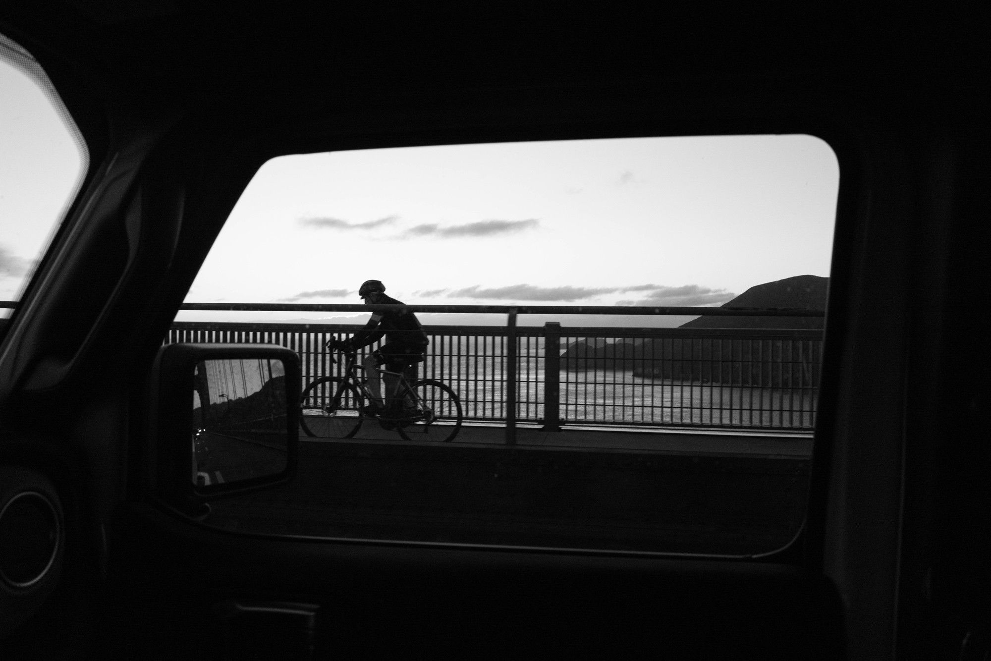 A black and white photo of a bike rider on a bridge framed through a vehicle’s passenger-side window. 