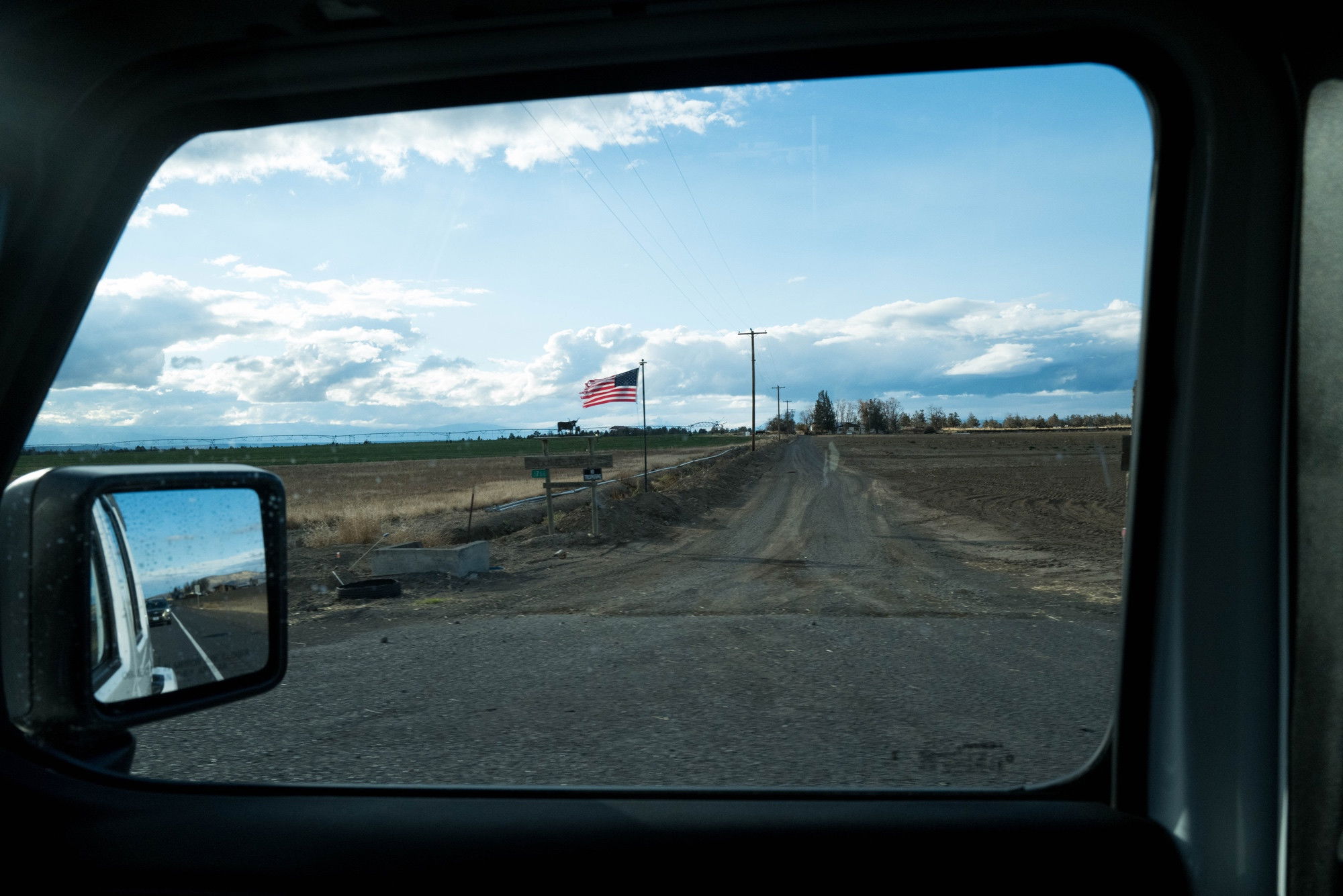 A color photo of an American flag by a dirt road framed through a vehicle’s passenger-side window. 