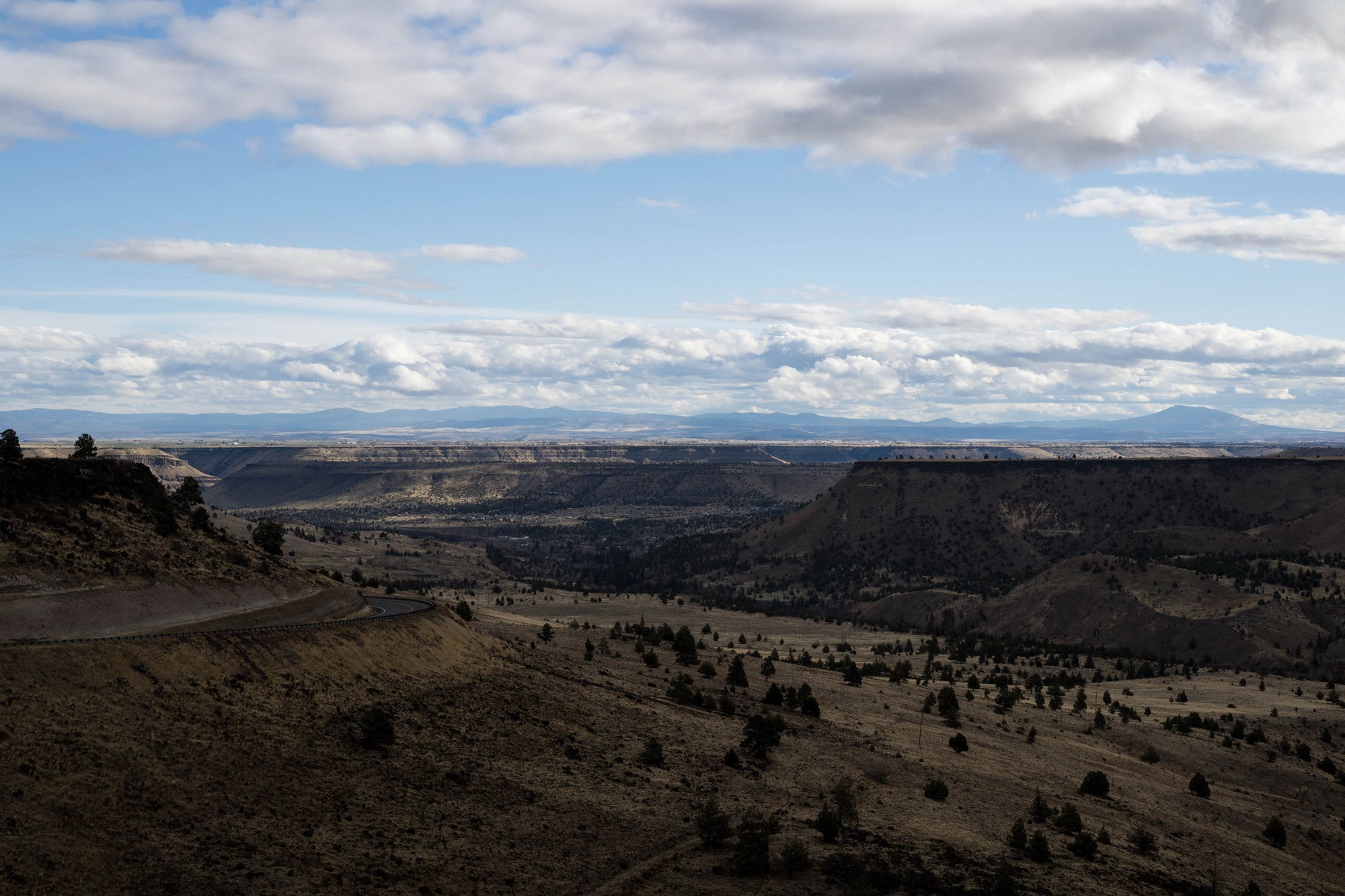 Panoramic view of high desert canyon with layered cliffs and mesas, winding road on left, scattered junipers, distant mountains under blue sky with clouds.