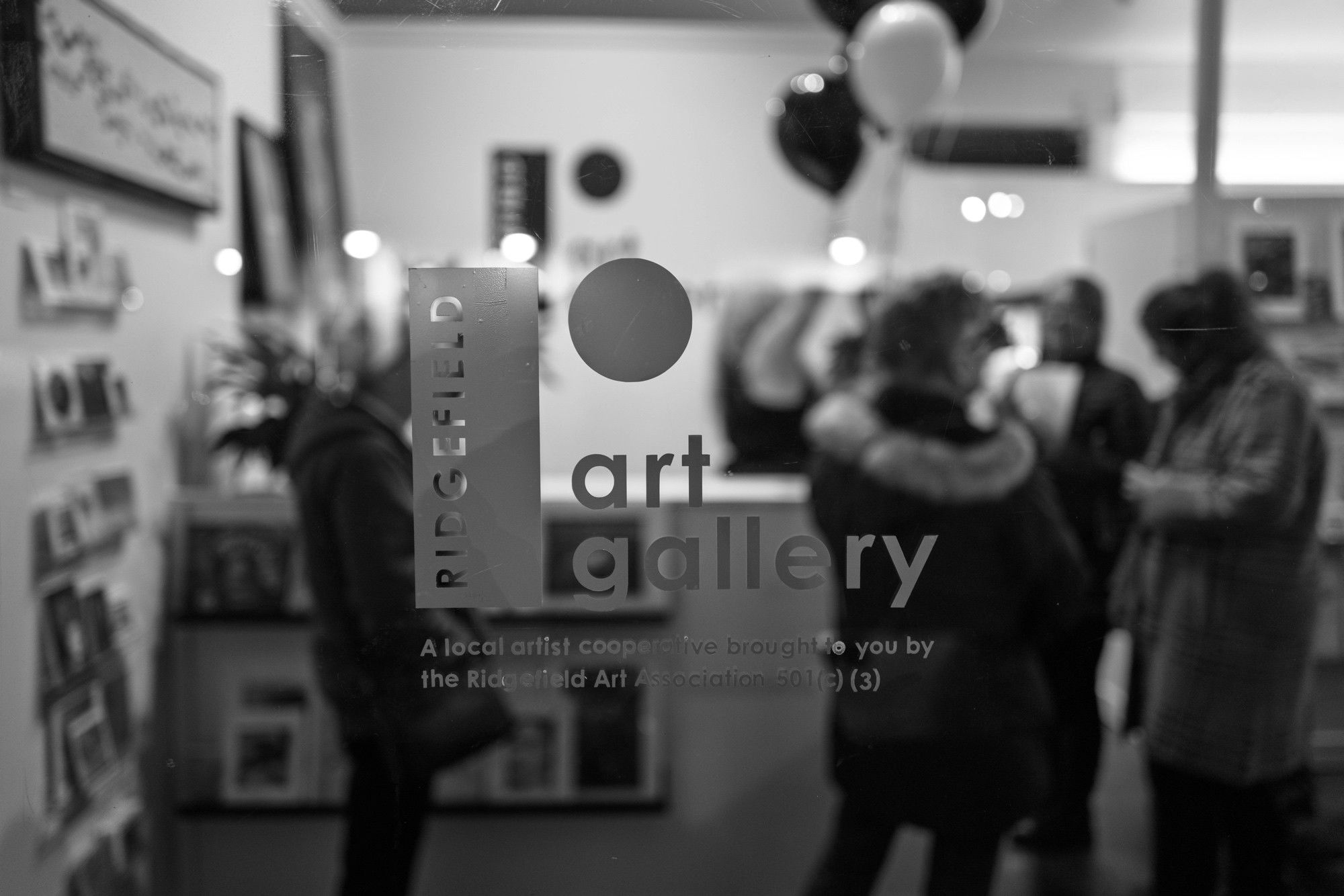 Window sign reading “RIDGEFIELD art gallery” with tagline “A local artist cooperative brought to you by the Ridgefield Art Association 501(c)(3)” with blurred visitors and balloons inside.